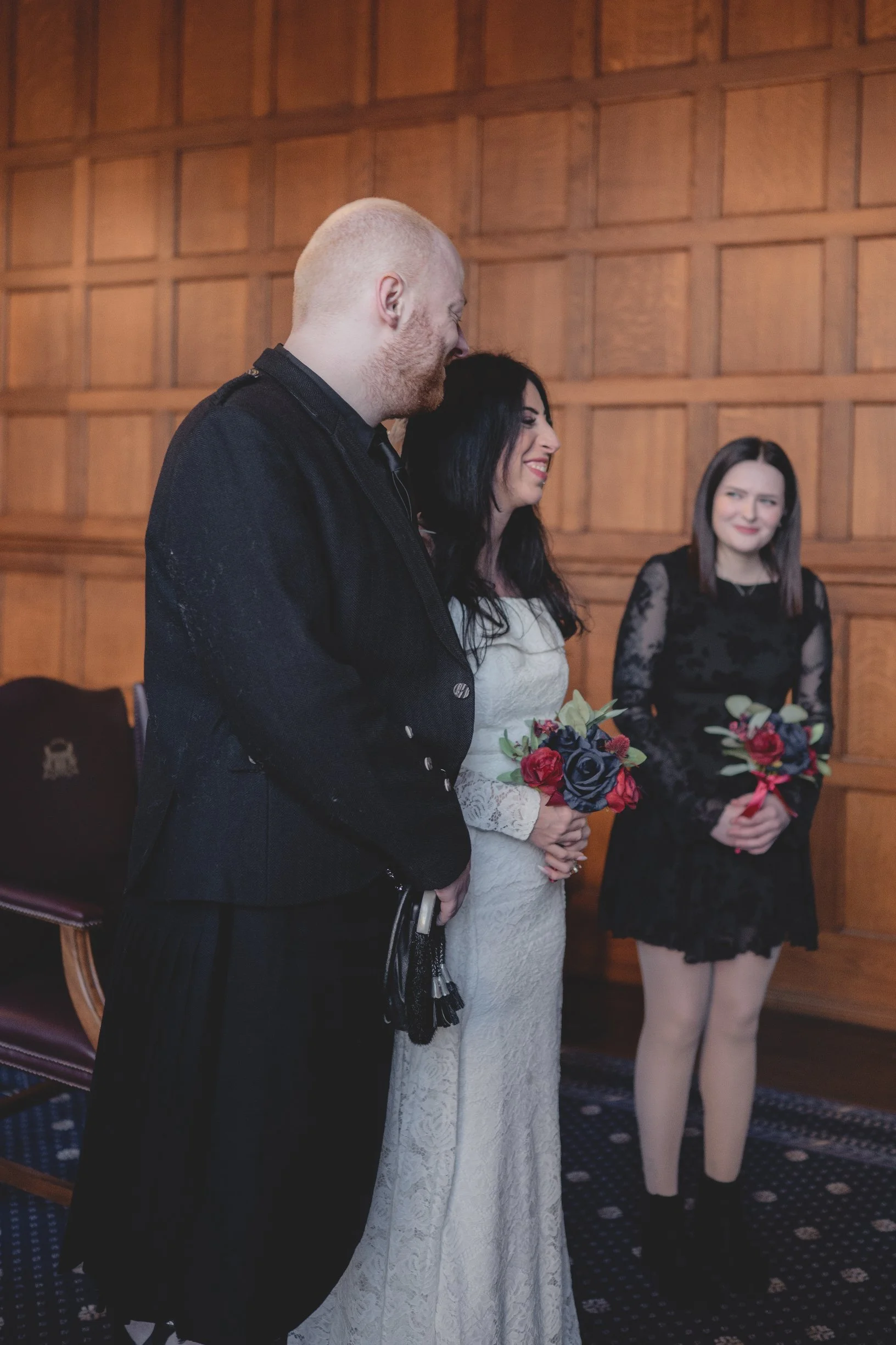 A wedding ceremony with a groom, bride, and a bridesmaid, standing in a wood-paneled room. The groom is wearing a traditional Scottish kilt, the bride has dark hair and is smiling while holding a bouquet, and the bridesmaid has dark hair, wears a bla