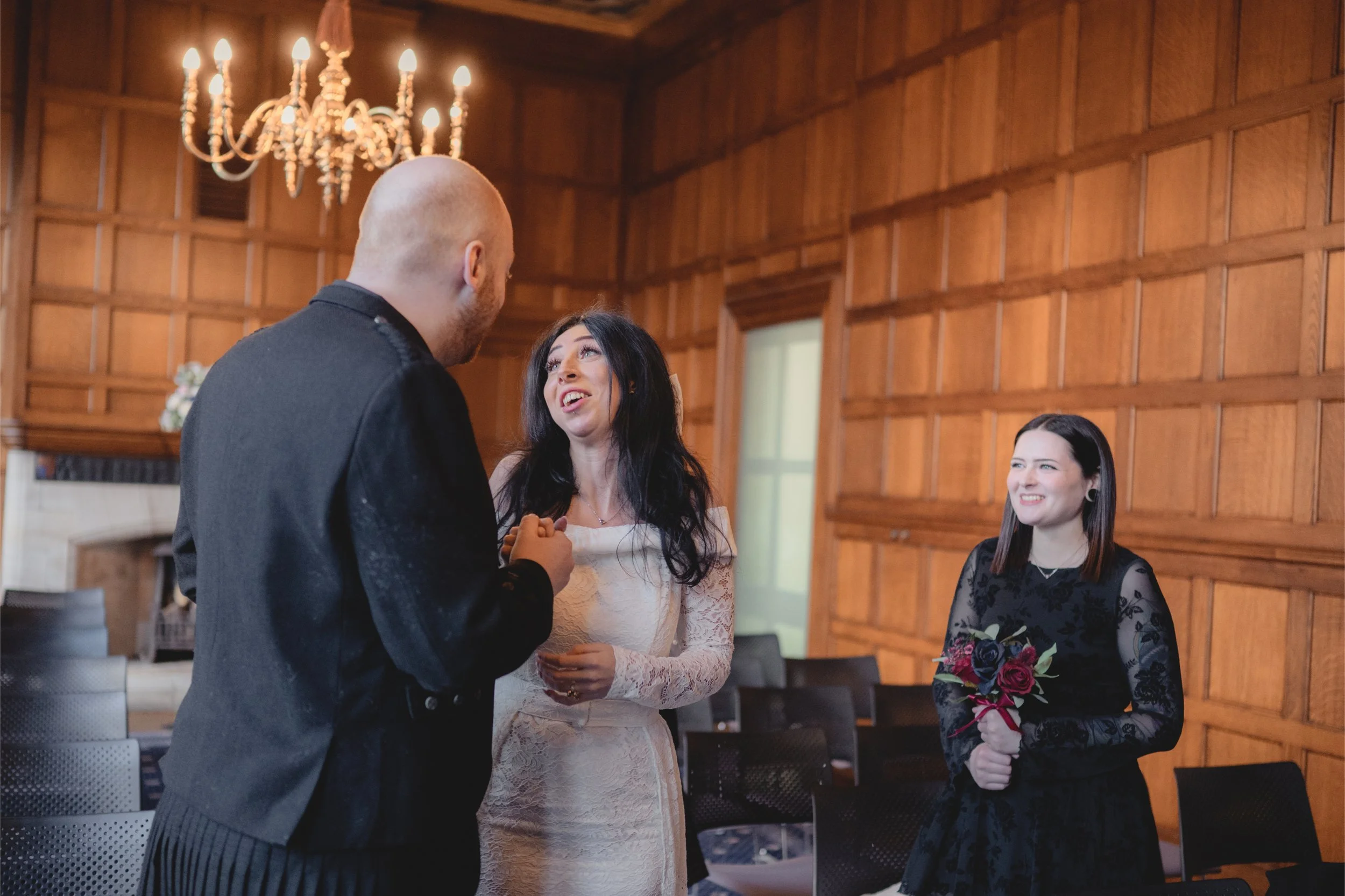 A man and woman are holding hands and talking while a woman holding a bouquet watches and smiles at them in a room with wood-paneled walls and a chandelier.