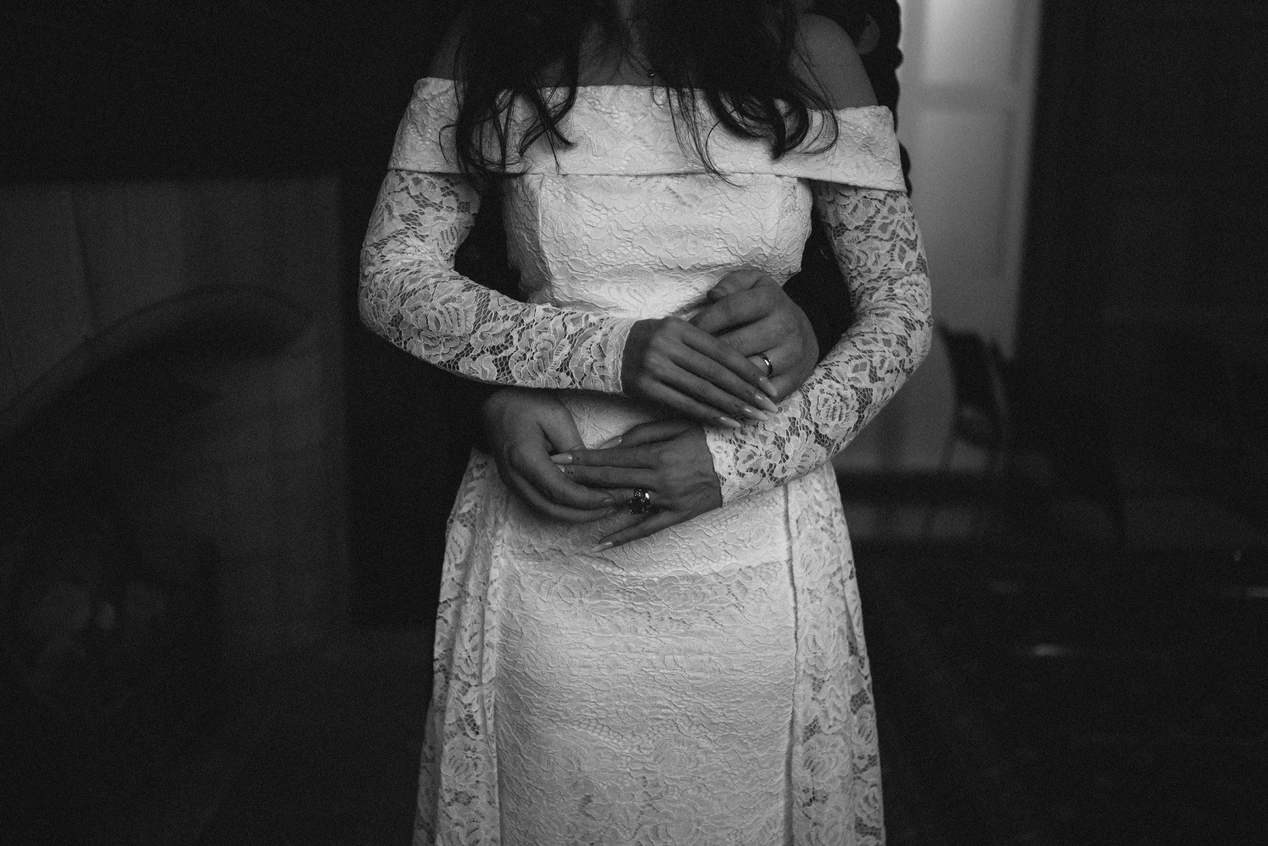 A woman in a white lace off-the-shoulder gown standing with her hands clasped in front of her, wearing rings on her fingers, in a dimly lit room.