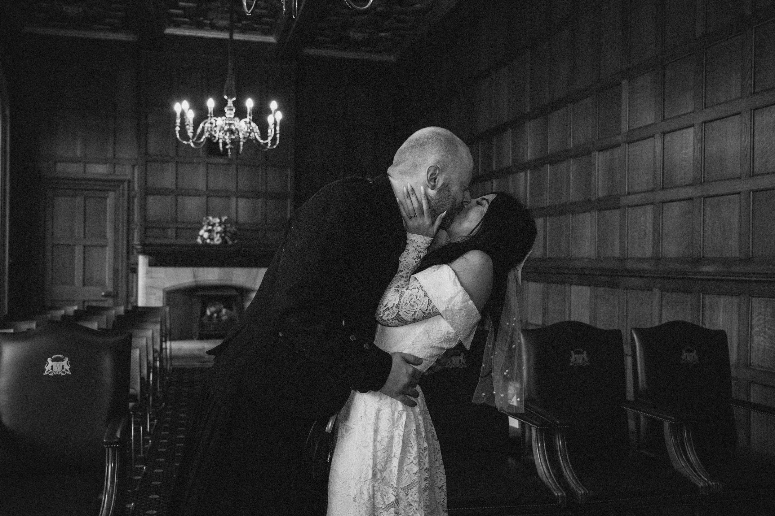 A man and woman kissing passionately in a dimly lit room with wooden paneling, chandelier, and empty chairs.