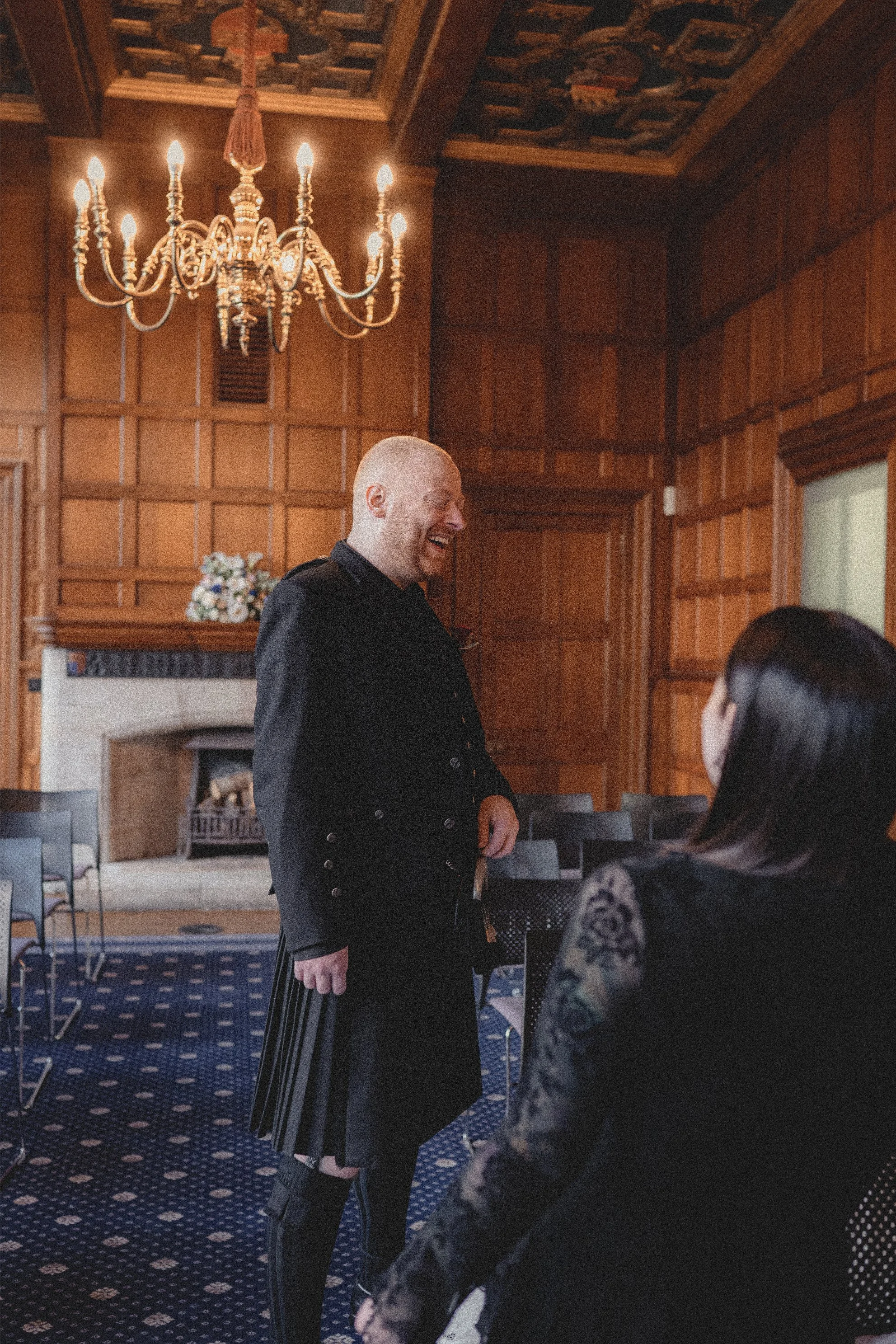 A man dressed in traditional Scottish attire, including a kilt, laughing and engaging with a woman in a lace dress inside a wood-paneled room with a chandelier and a fireplace.