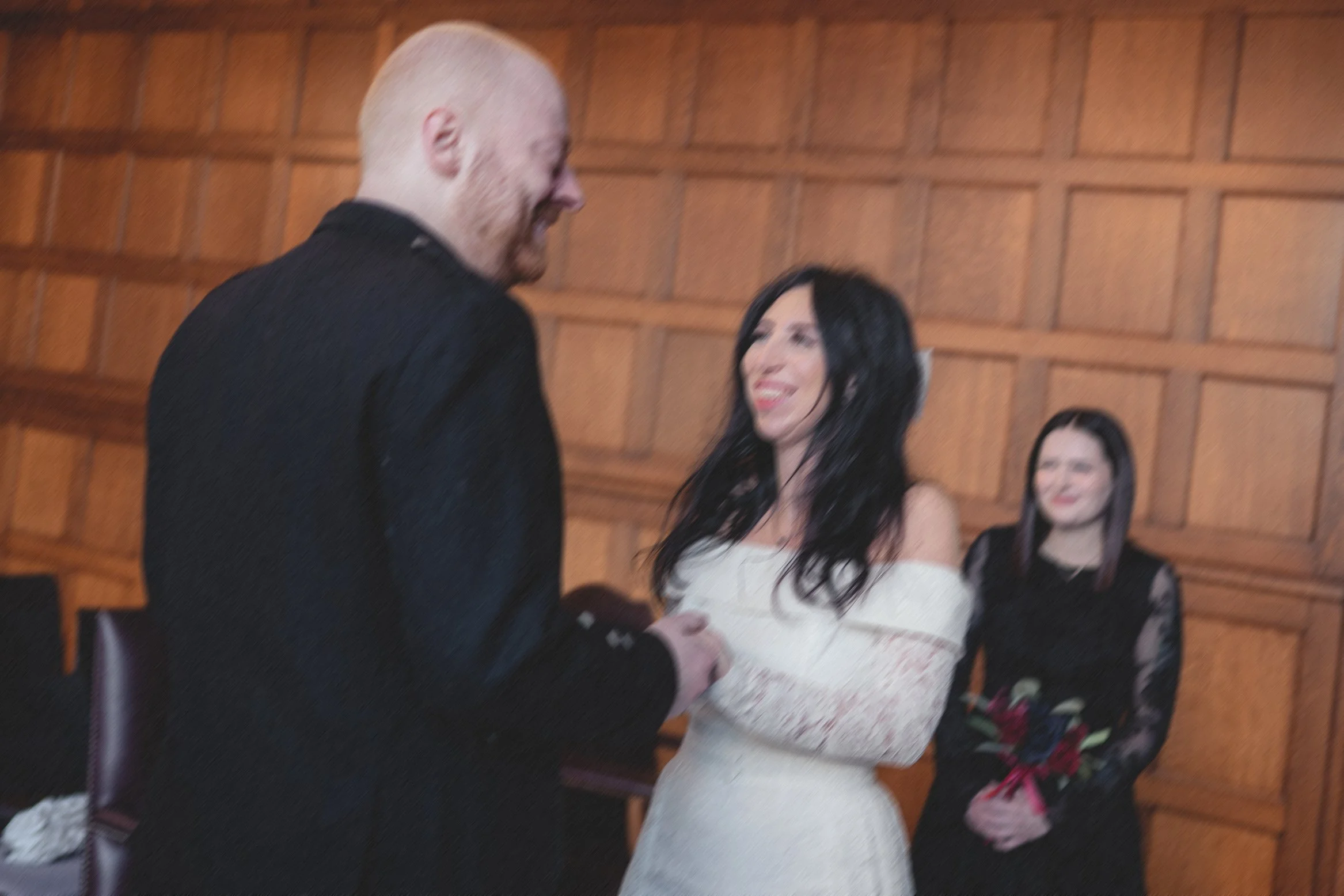 A woman in a white off-shoulder dress and a man in a black suit are exchanging vows and holding hands during a wedding ceremony, with another woman in a black lace dress standing in the background.