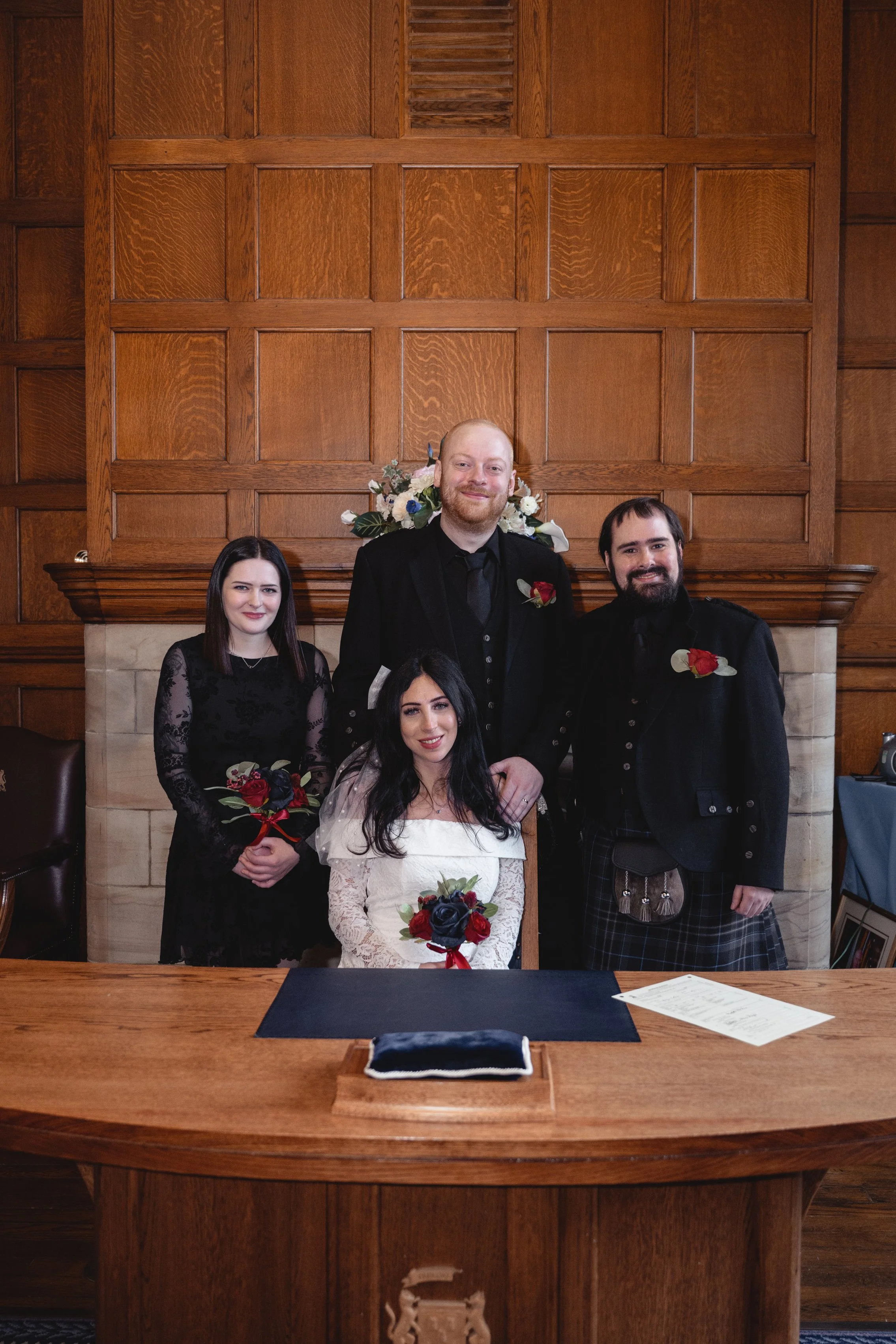 Group of five people at a wedding ceremony, standing behind a wooden table with a document and a cloth, in front of a wooden paneled wall and fireplace with flowers. The bride is seated in front, wearing a white lace dress and holding a bouquet. The 