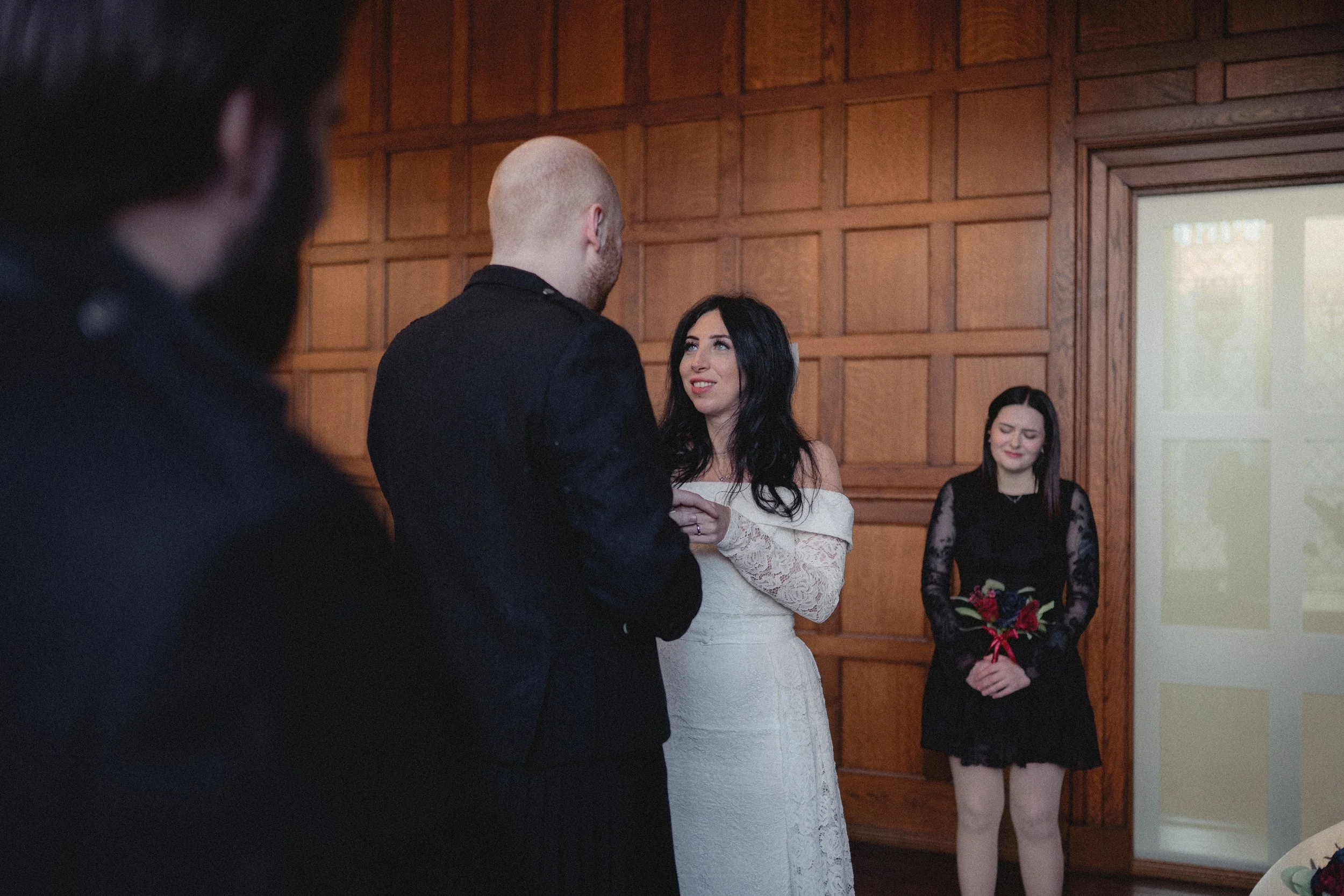 A wedding ceremony with a bride dressed in white and a groom in black standing in front of a woman holding a bouquet, with another woman smiling in the background.
