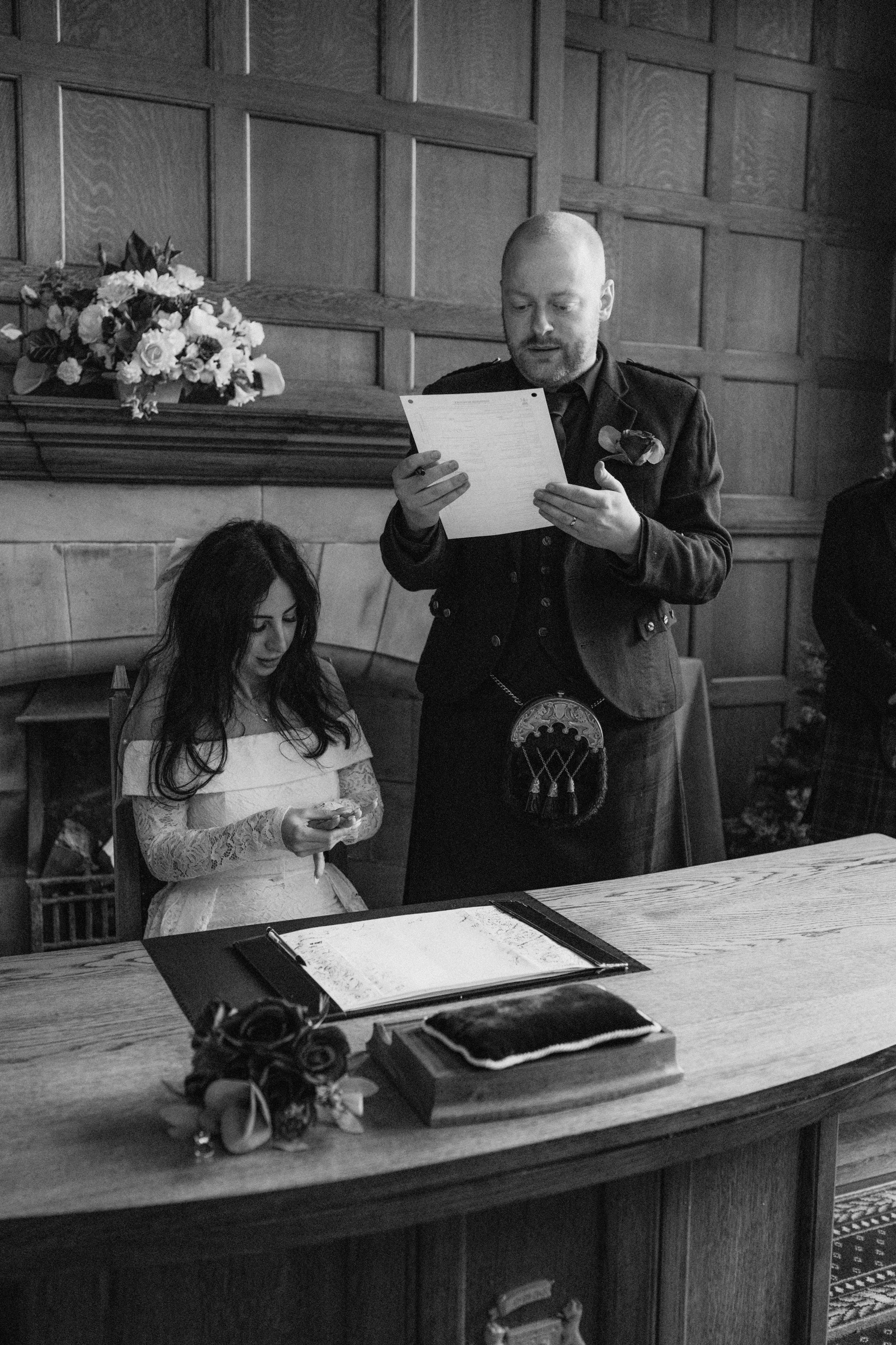 A man, dressed in traditional Scottish attire with a kilt and sporran, appears to be reading vows or a speech during a wedding ceremony, standing next to a young girl in a white dress who is holding a ring, in a wood-paneled room with a bouquet of fl