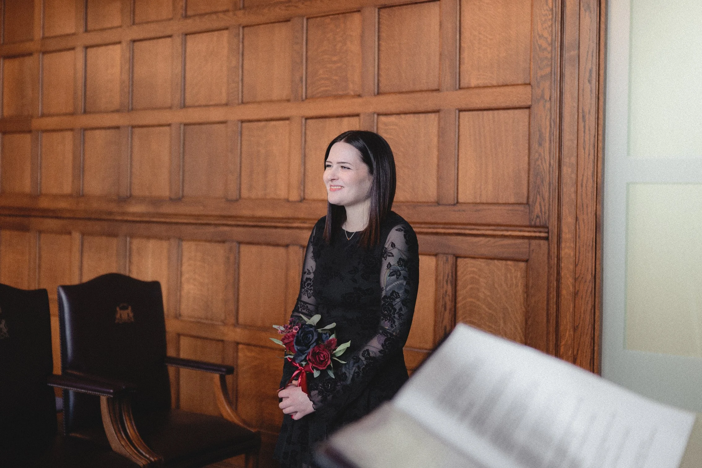 A woman with dark brown hair, wearing a black lace dress, is standing in a wooden-paneled room, holding a bouquet of red and black flowers, and smiling.