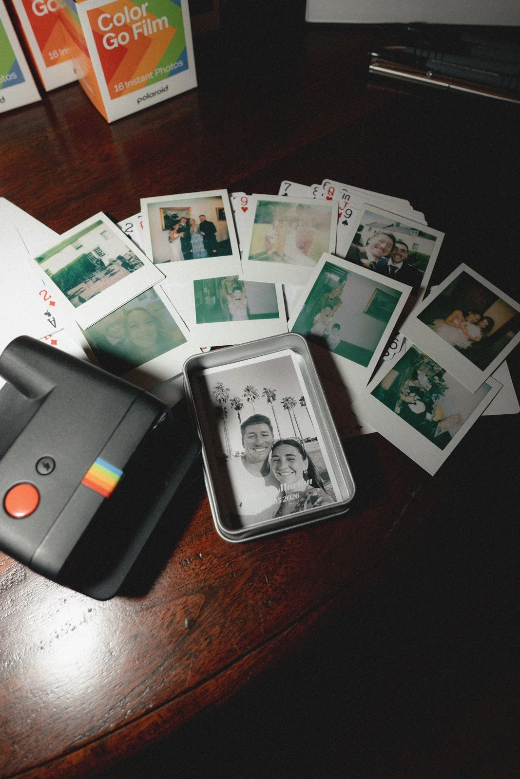 Collection of printed photographs, instant film pictures, and a camera on a wooden surface. The photos include people, outdoor scenes, and a black-and-white image of a smiling couple with palm trees in the background.
