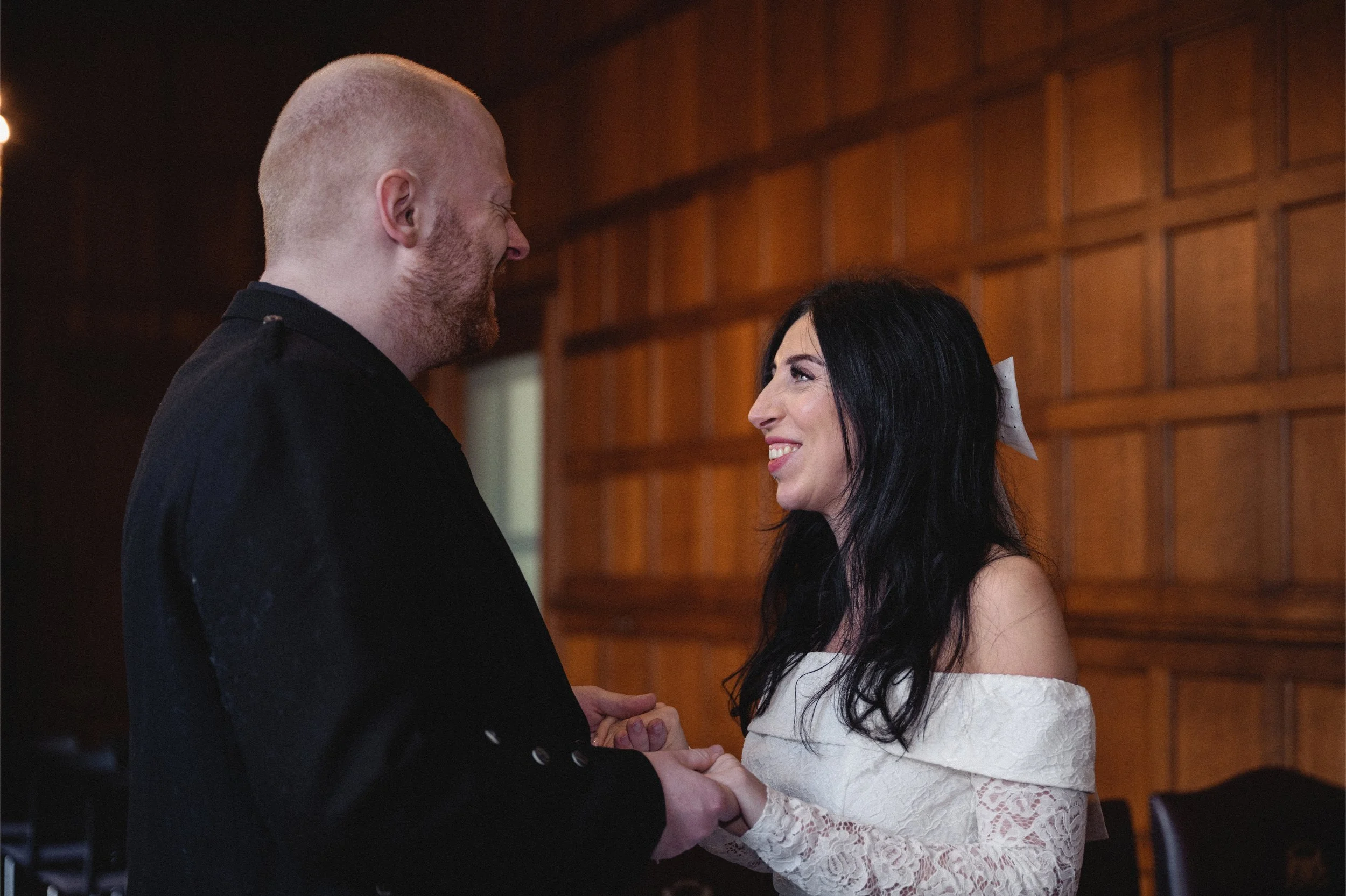 A man and woman holding hands and smiling at each other in a warmly lit room with wooden paneling.