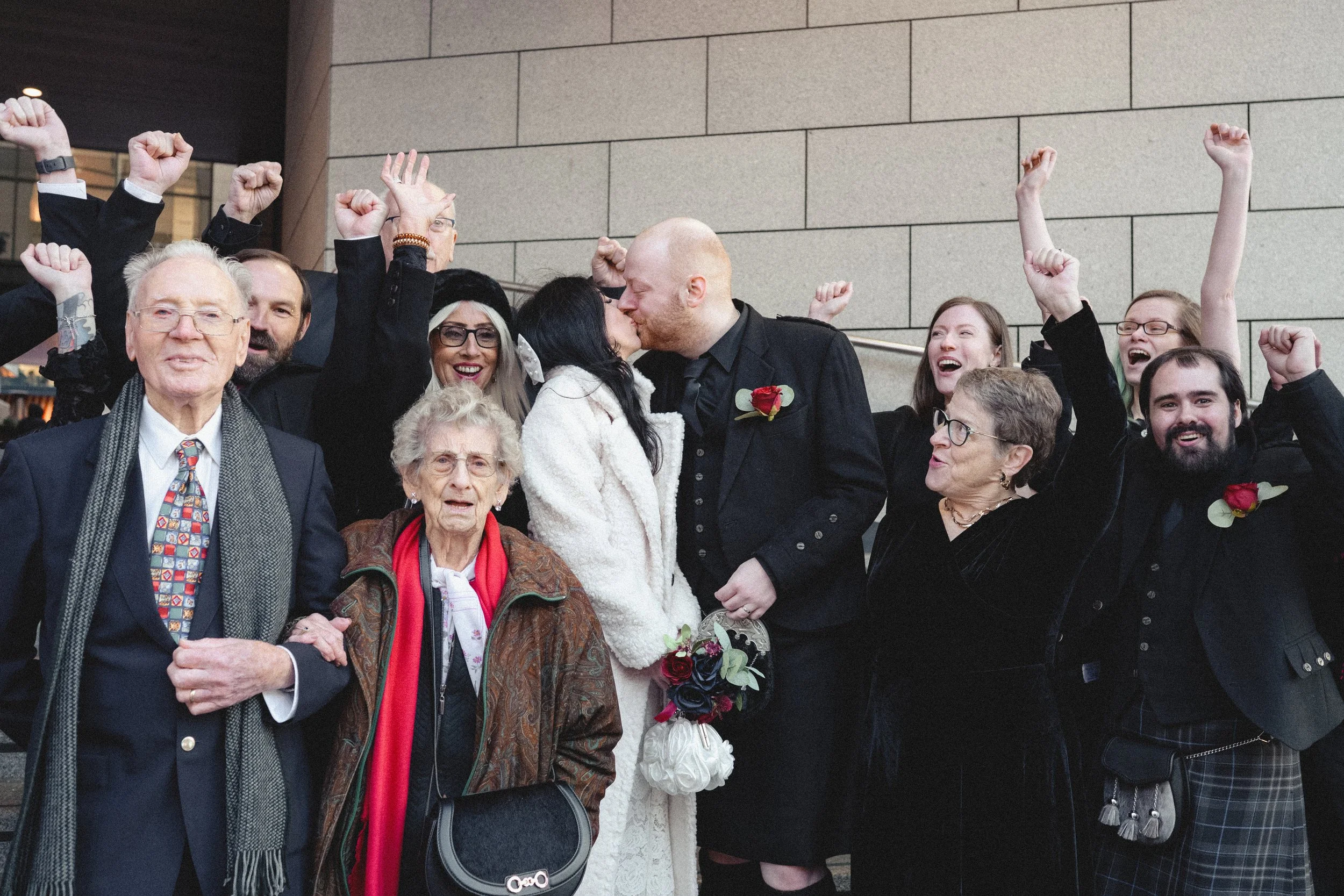 A group of people celebrating, with some raising their fists in the air, one couple kissing in front of the group, and others smiling and showing excitement during a special occasion, possibly a wedding.