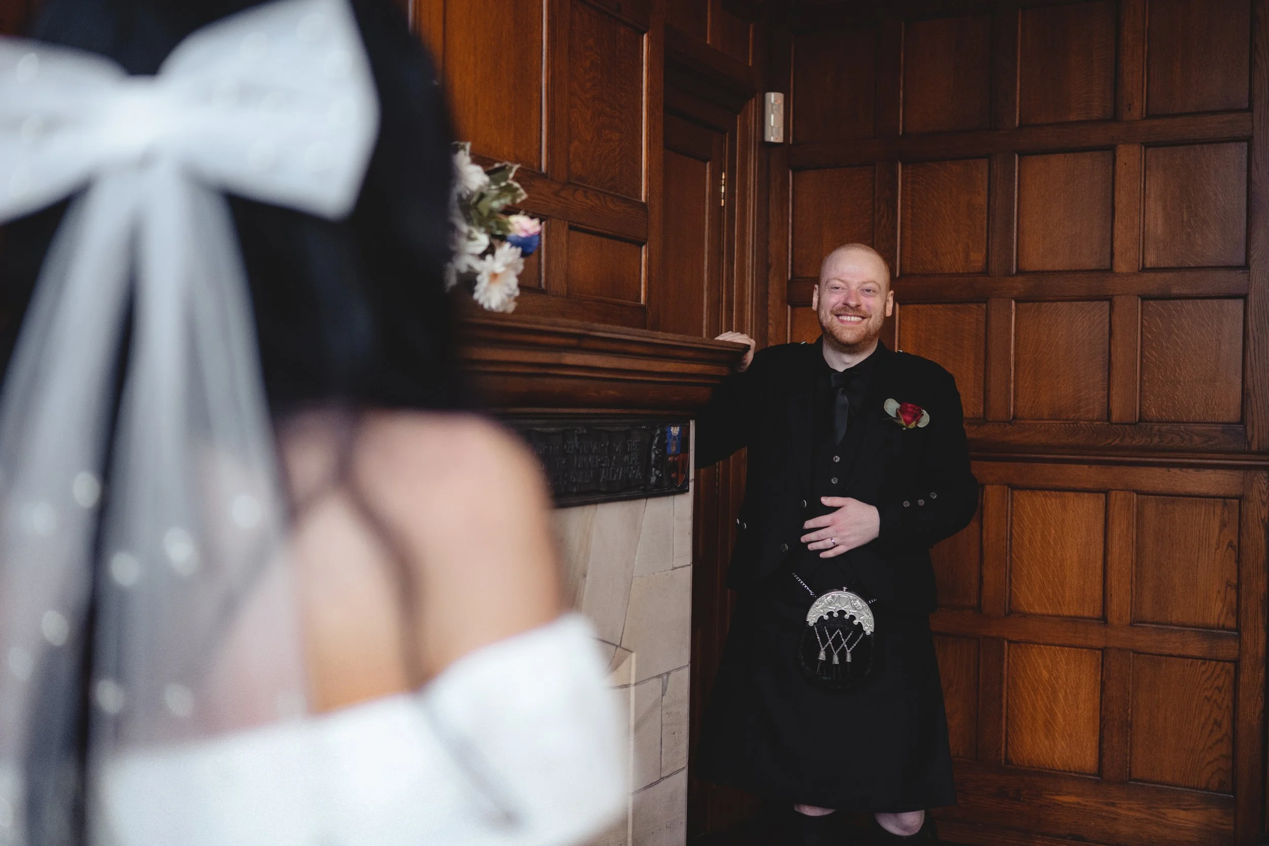 A smiling man dressed in formal black attire standing in a wooden-paneled room, looking at a woman with blurred dark hair in the foreground, wearing a white dress with a voluminous skirt and a large white bow in her hair.