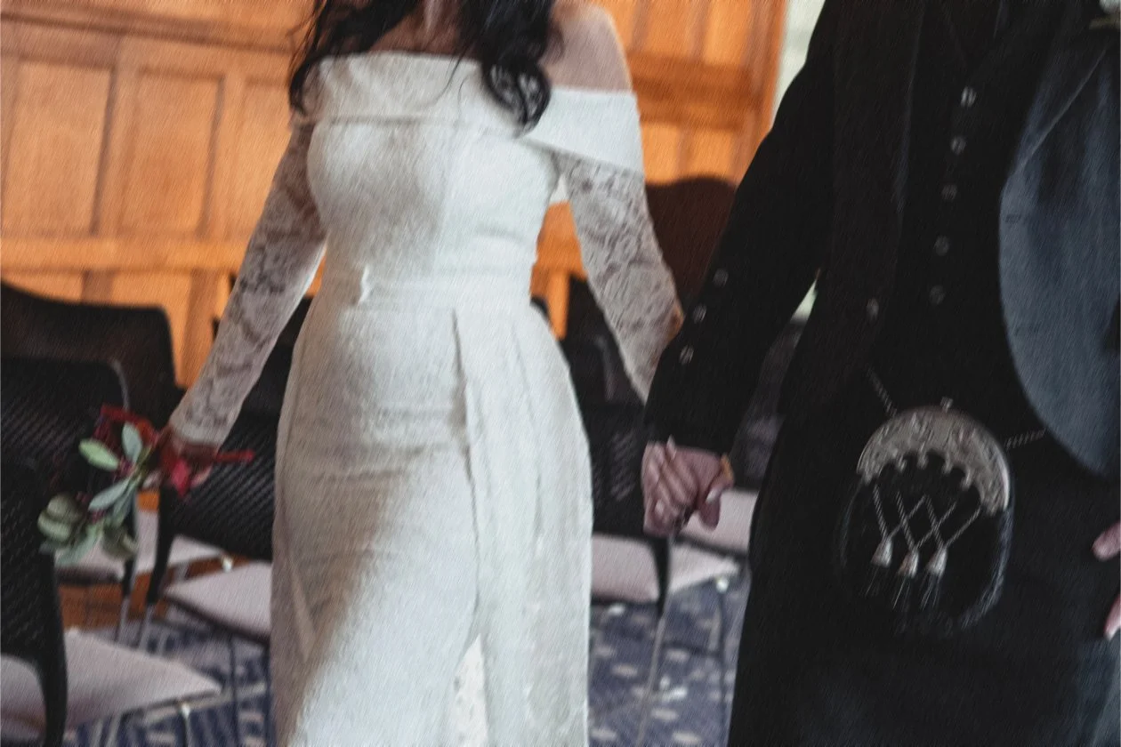 A couple holding hands, with the woman in a white wedding dress and the man in a black tuxedo, at a wedding ceremony.