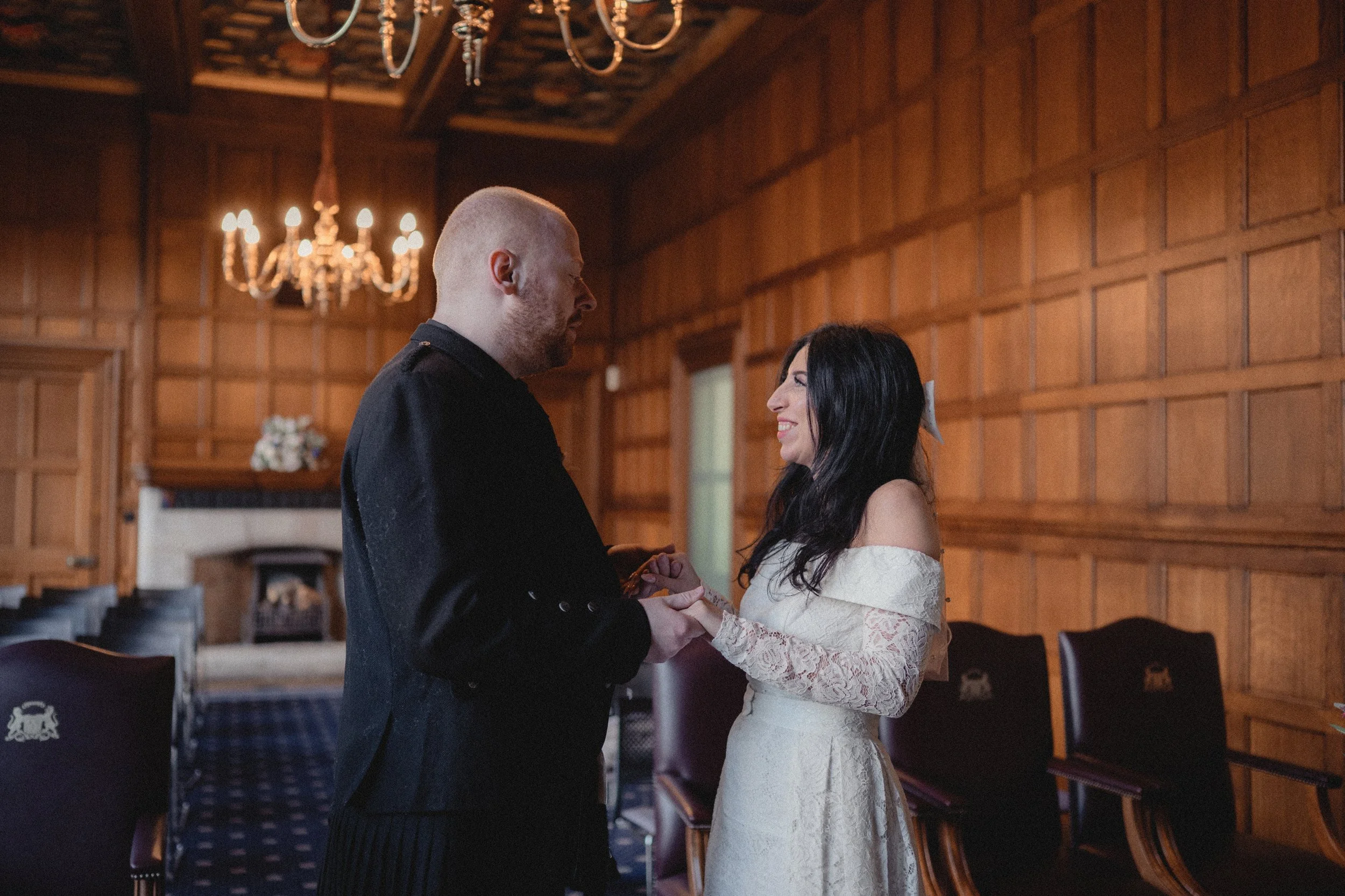 A couple holding hands and smiling at each other in a wood-paneled room with chandeliers, possibly during a wedding or romantic event.