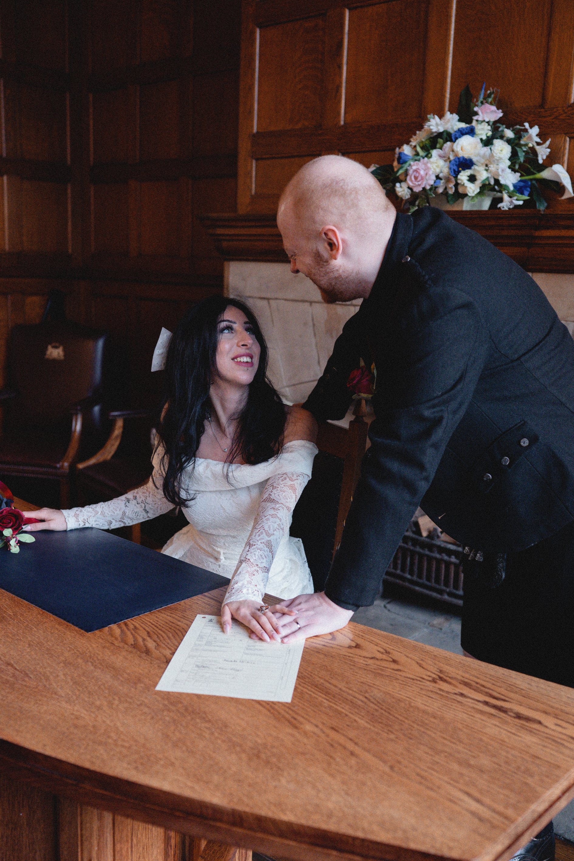 A woman in a white lace off-the-shoulder dress and a man with a bald head and black attire share a moment at a wedding or ceremony, with a floral arrangement in the background.