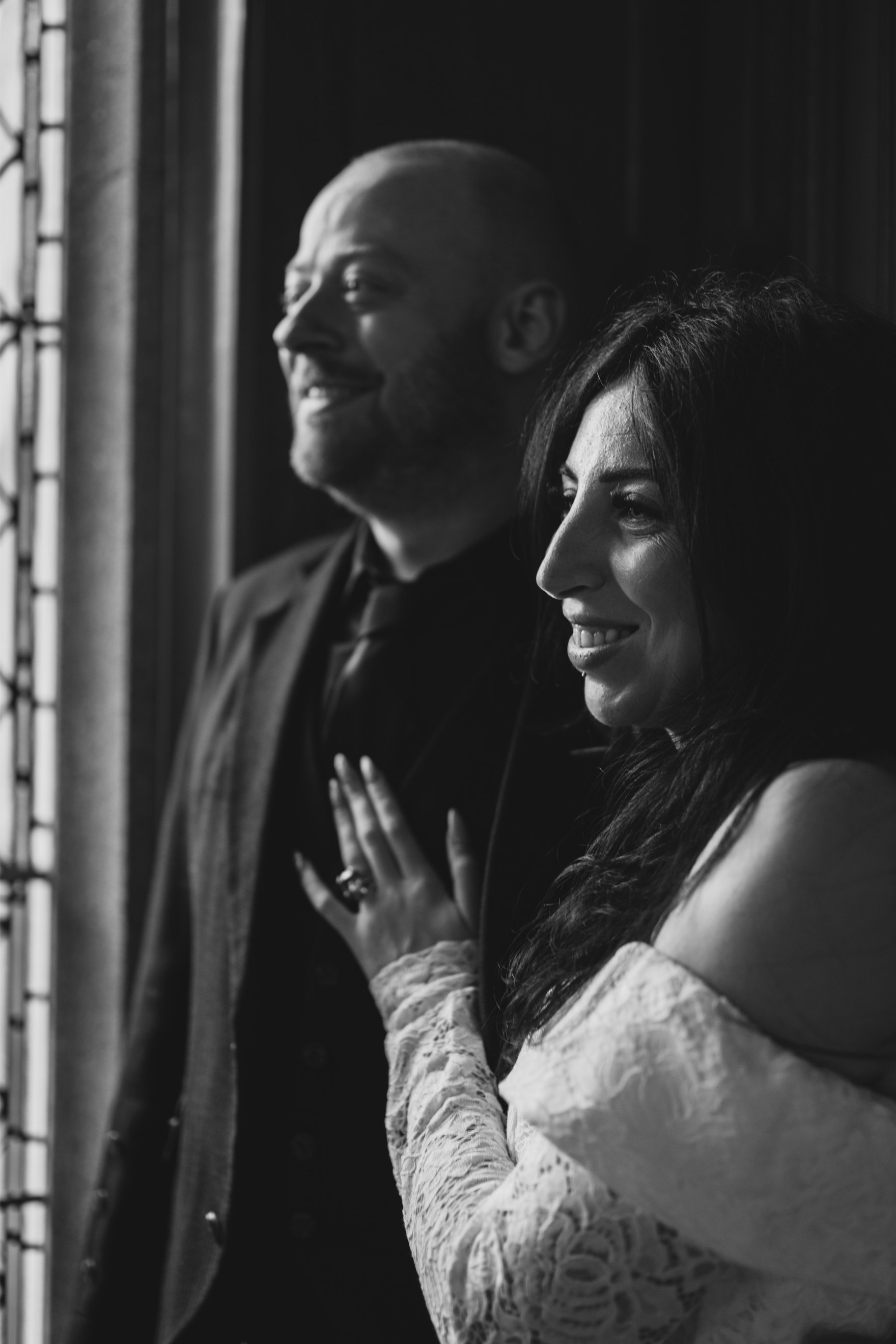 Black and white photo of a smiling man and woman standing close together near a window, with the woman holding her hand near her chest and wearing an off-shoulder lace dress.