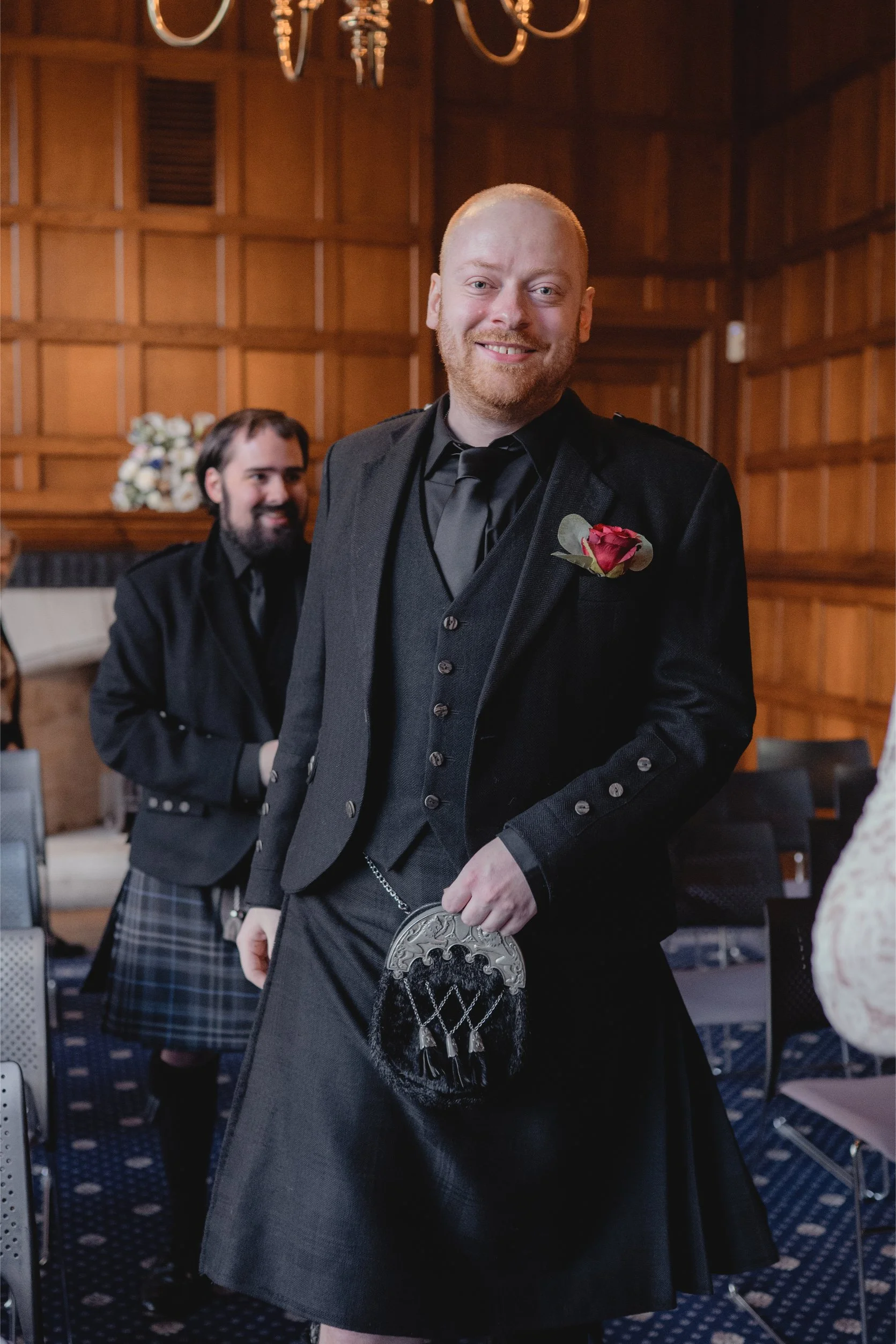 A man in traditional Scottish attire, including a kilt, standing indoors with a smile, holding a decorative sporran. Another man in similar attire stands in the background, also smiling. The setting appears to be a formal event in a wood-paneled room