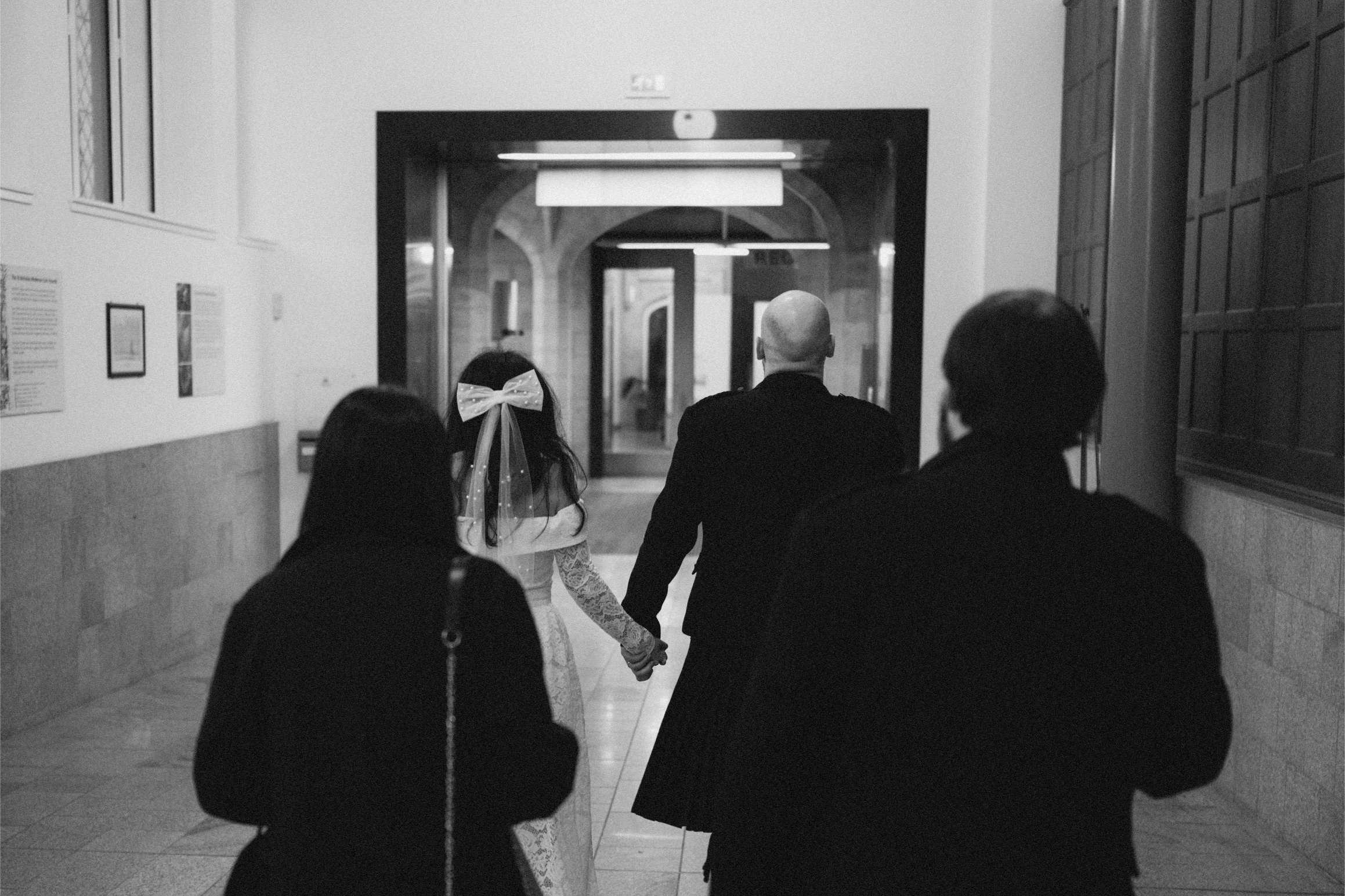 Black and white photo of a couple walking hand in hand, surrounded by two other people, through a hallway leading to a building entrance.