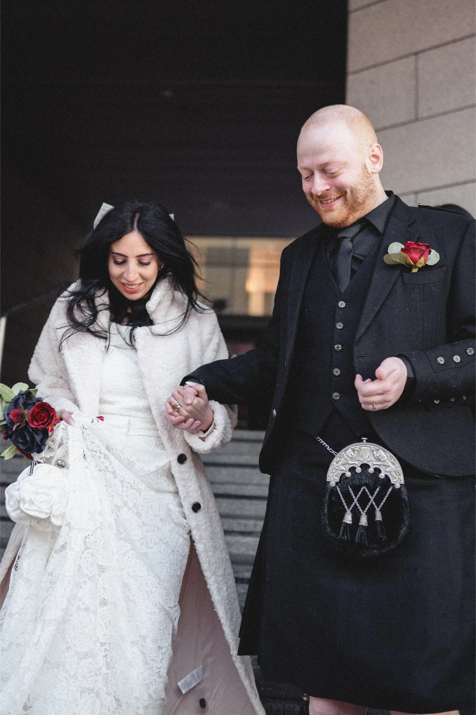 A bride and groom holding hands after a wedding ceremony, standing outdoors on steps, dressed in wedding attire, smiling.