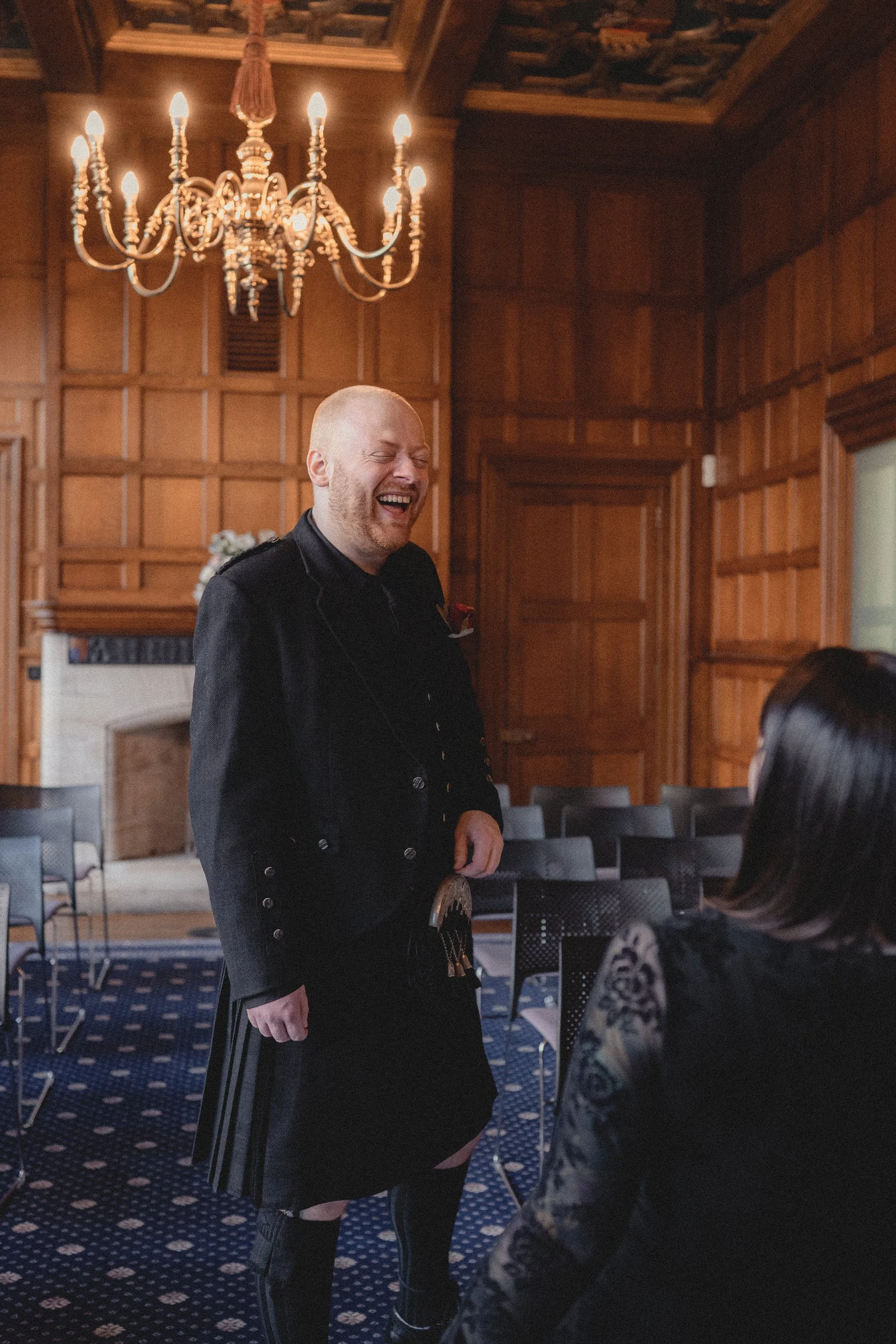 A man dressed in a kilt and black jacket laughing with a woman sitting in front of him in a wood-paneled room with a chandelier overhead.
