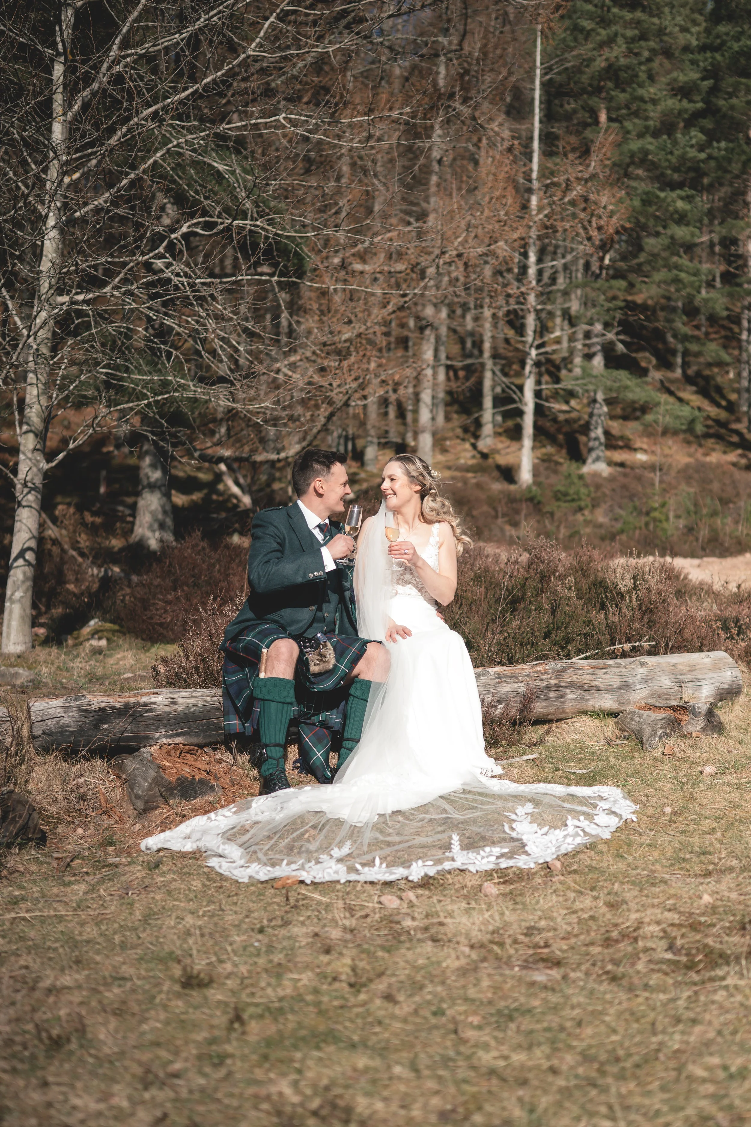 A bride and groom sitting on a log outdoors, toasting with champagne glasses, surrounded by trees and natural scenery.