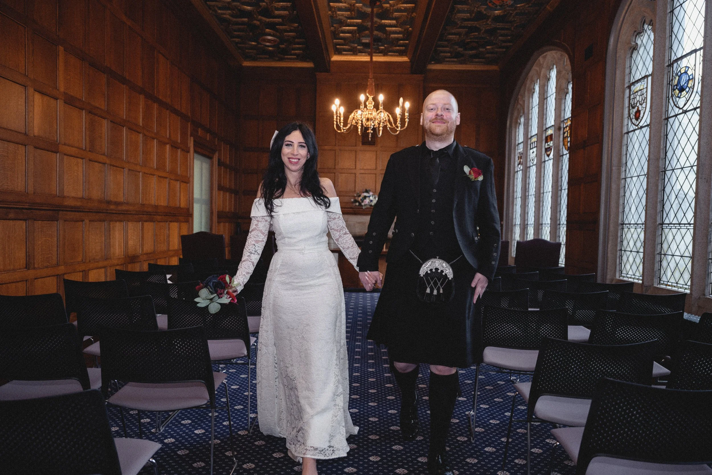 A bride and groom holding hands and walking inside a wood-paneled room with stained glass windows and a chandelier, dressed in wedding attire.