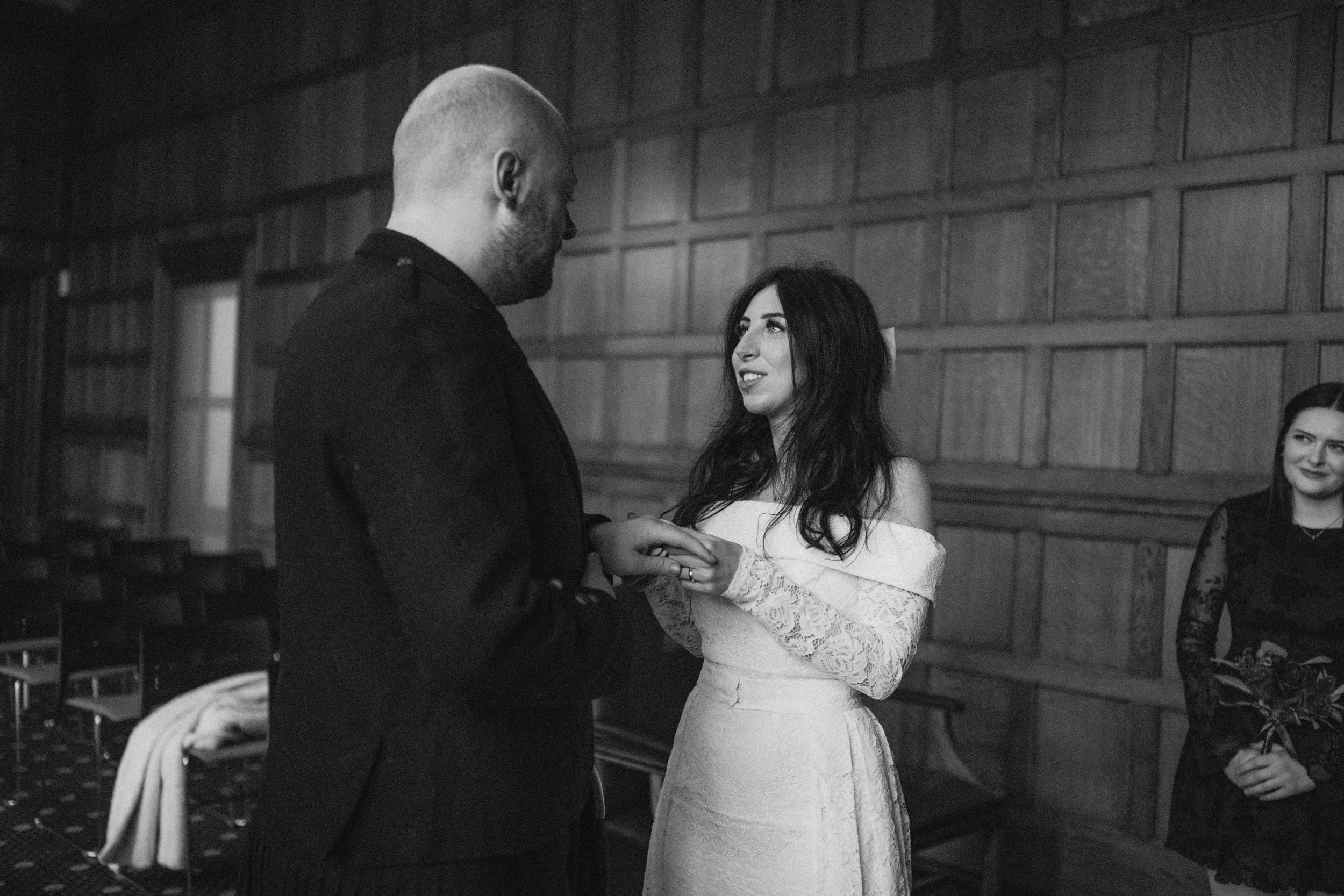 A man and woman exchanging rings during a wedding ceremony, with a woman holding a bouquet standing in the background.