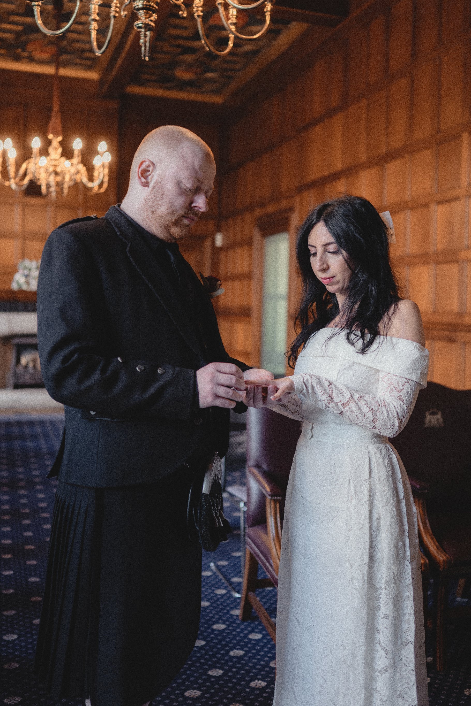 A man and woman exchanging rings during a wedding ceremony indoors with wood-paneled walls and chandeliers.