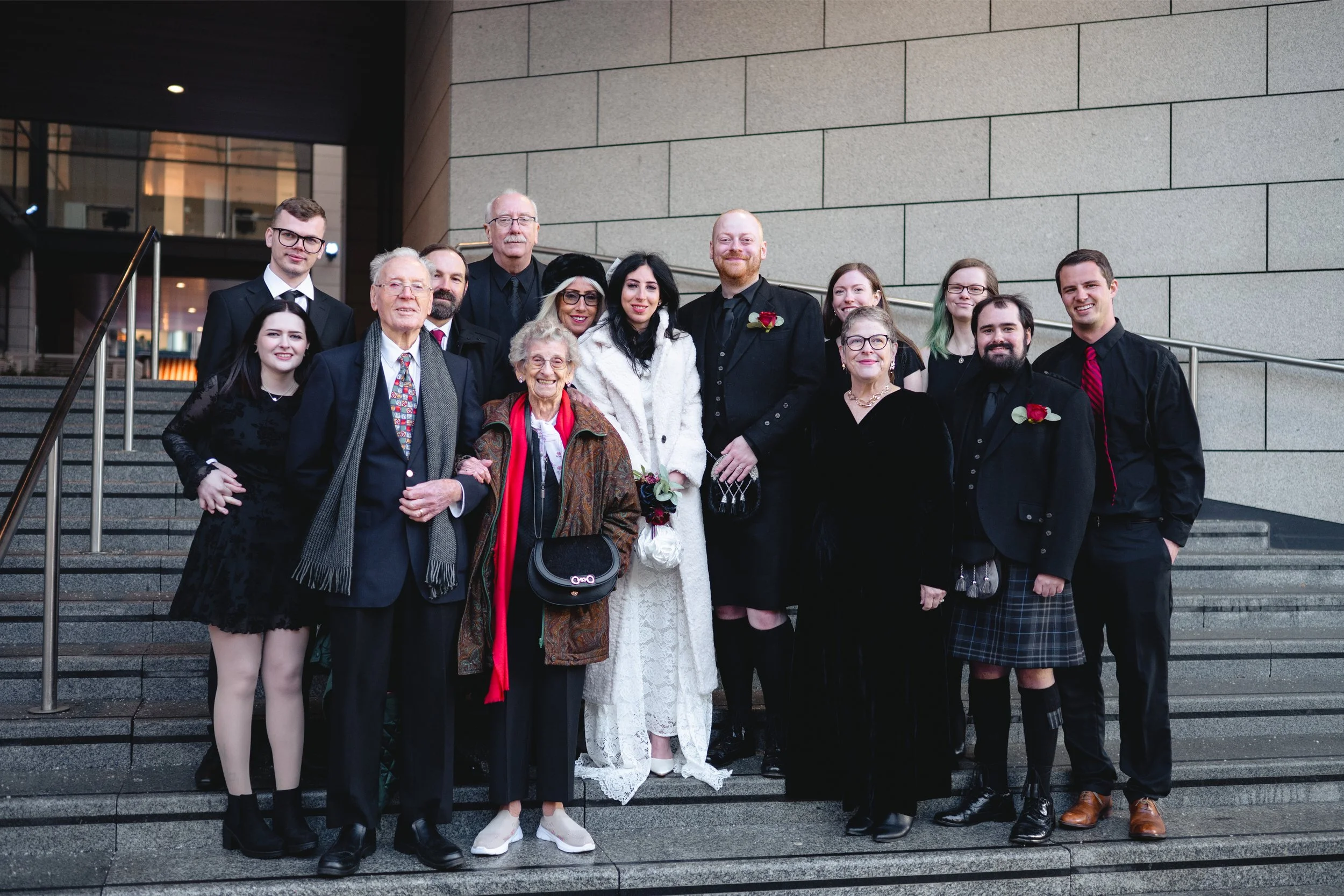 Group of people dressed in formal attire, standing on wedding venue steps. The bride is in white lace dress holding a bouquet. The groom in black suit with red boutonniere. Elderly couple, women in black dresses and glasses, men in suits and tartan k