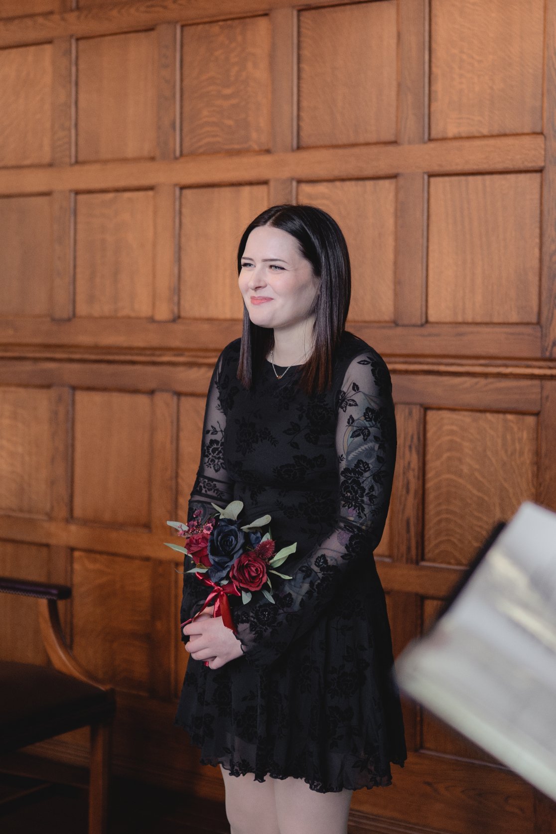 A young woman with black hair wearing a black floral dress holding a bouquet of red and dark purple roses with greenery, standing in front of a wooden wall.