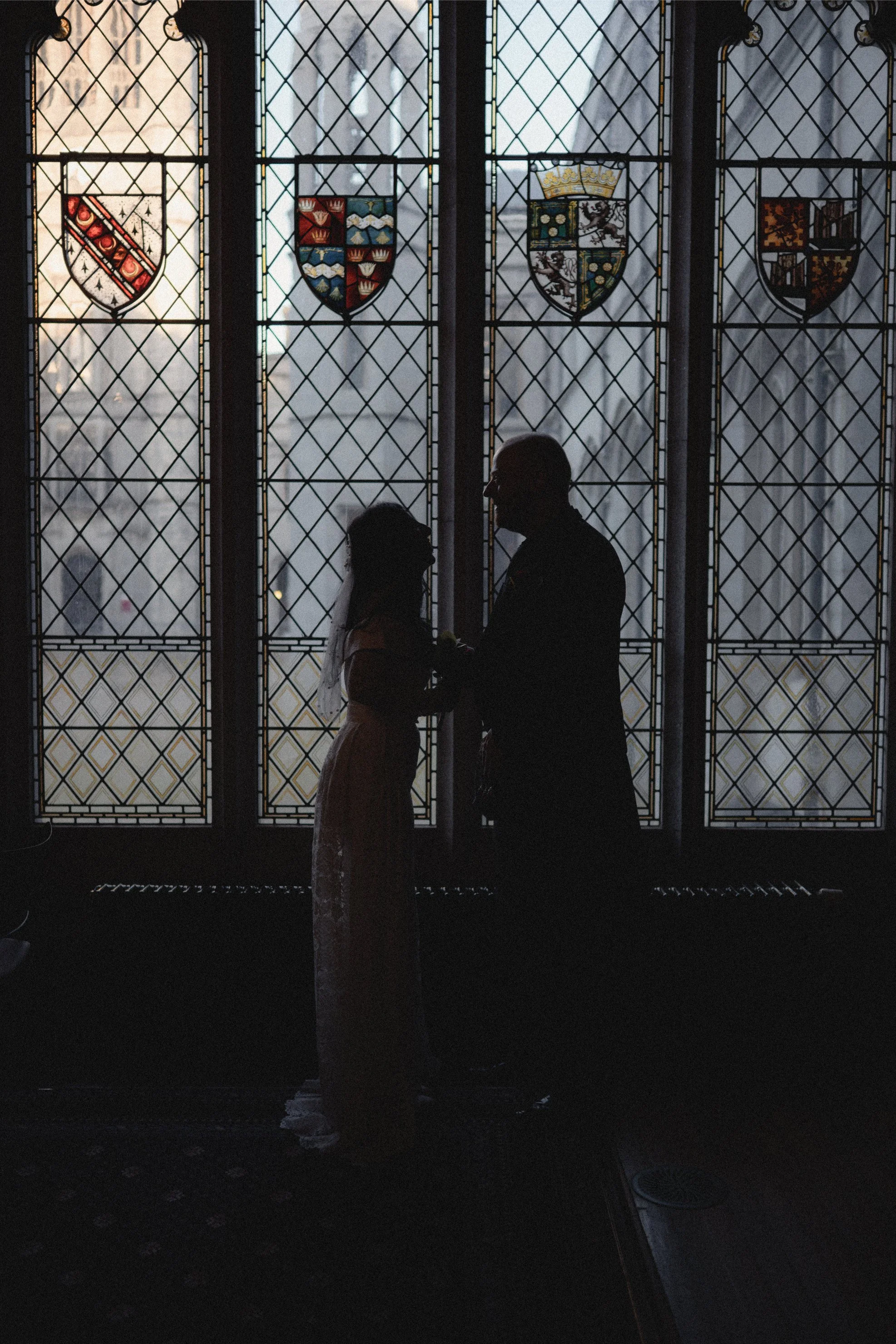 Silhouette of a bride and groom exchanging vows in front of stained glass windows with heraldic shields, inside a church.