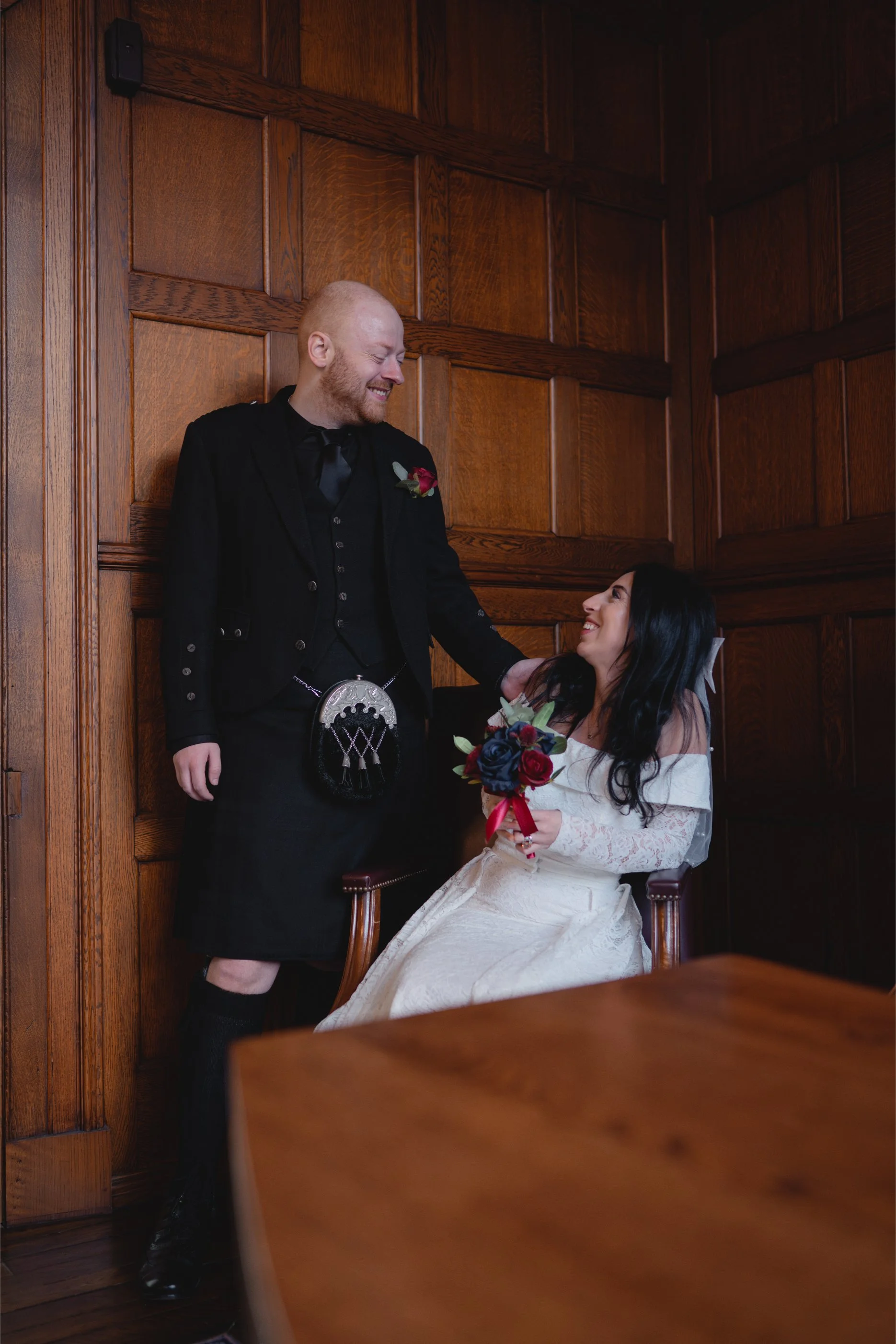 A groom and bride sharing a joyful moment indoors, with the groom leaning against a wooden wall and the bride sitting on a chair, holding a bouquet of flowers, both smiling at each other.