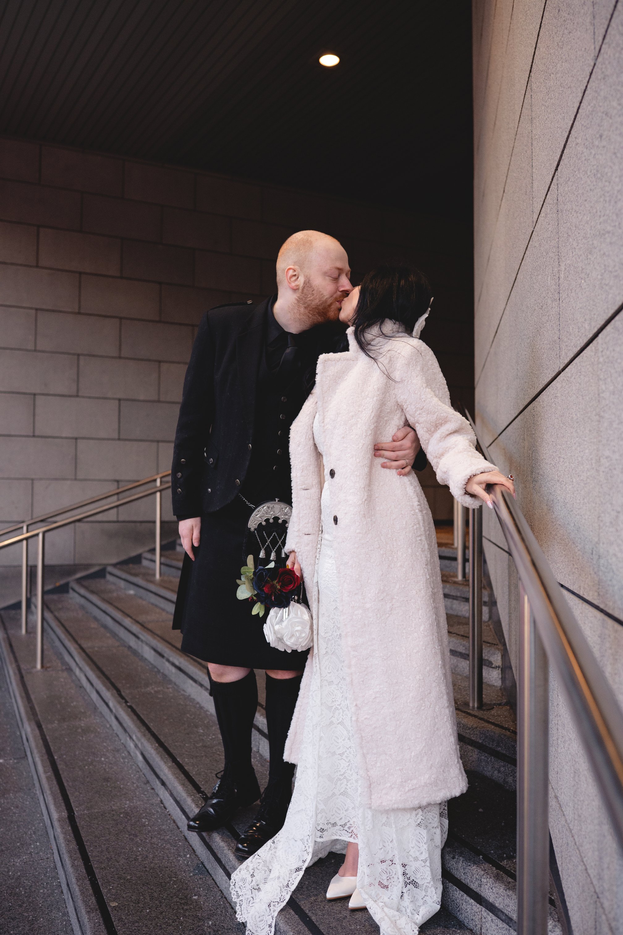 A couple sharing a kiss on a staircase, dressed in wedding attire with a brick and concrete wall background.