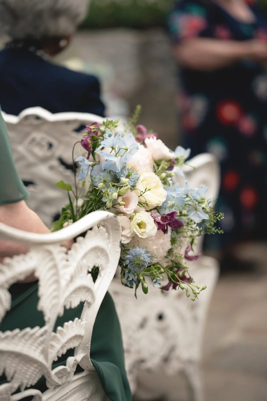 Close-up of a bouquet of white, blue, purple, and green flowers resting on a white ornate bench with people blurred in the background.