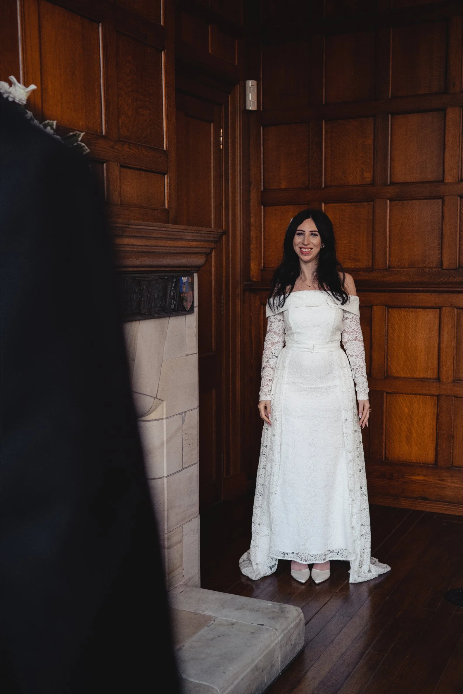 A woman in a white lace wedding dress standing in a room with wood-paneled walls, smiling.