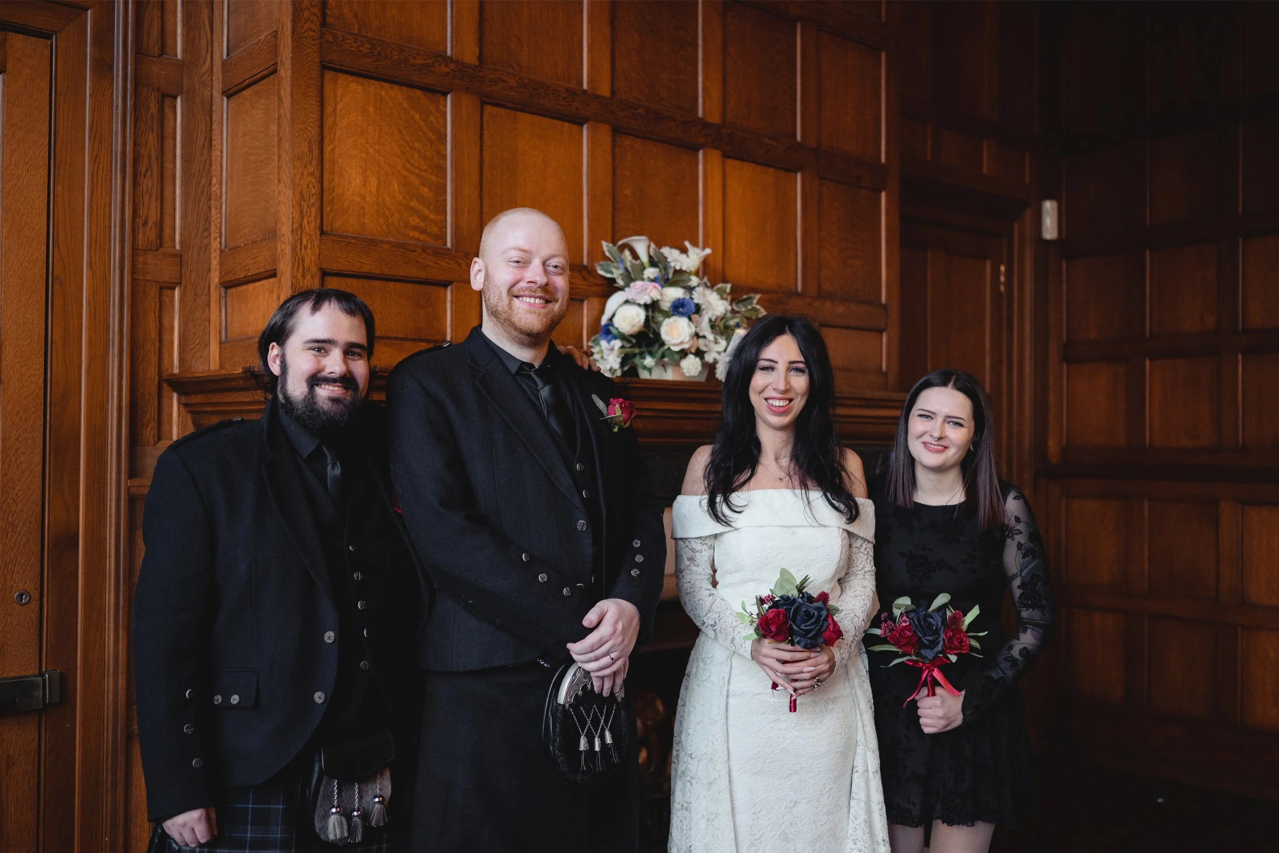 Four people smiling and standing in front of a fireplace with a floral arrangement on the mantel, in a room with wooden-paneled walls. Two women and two men, dressed formally, holding bouquets.