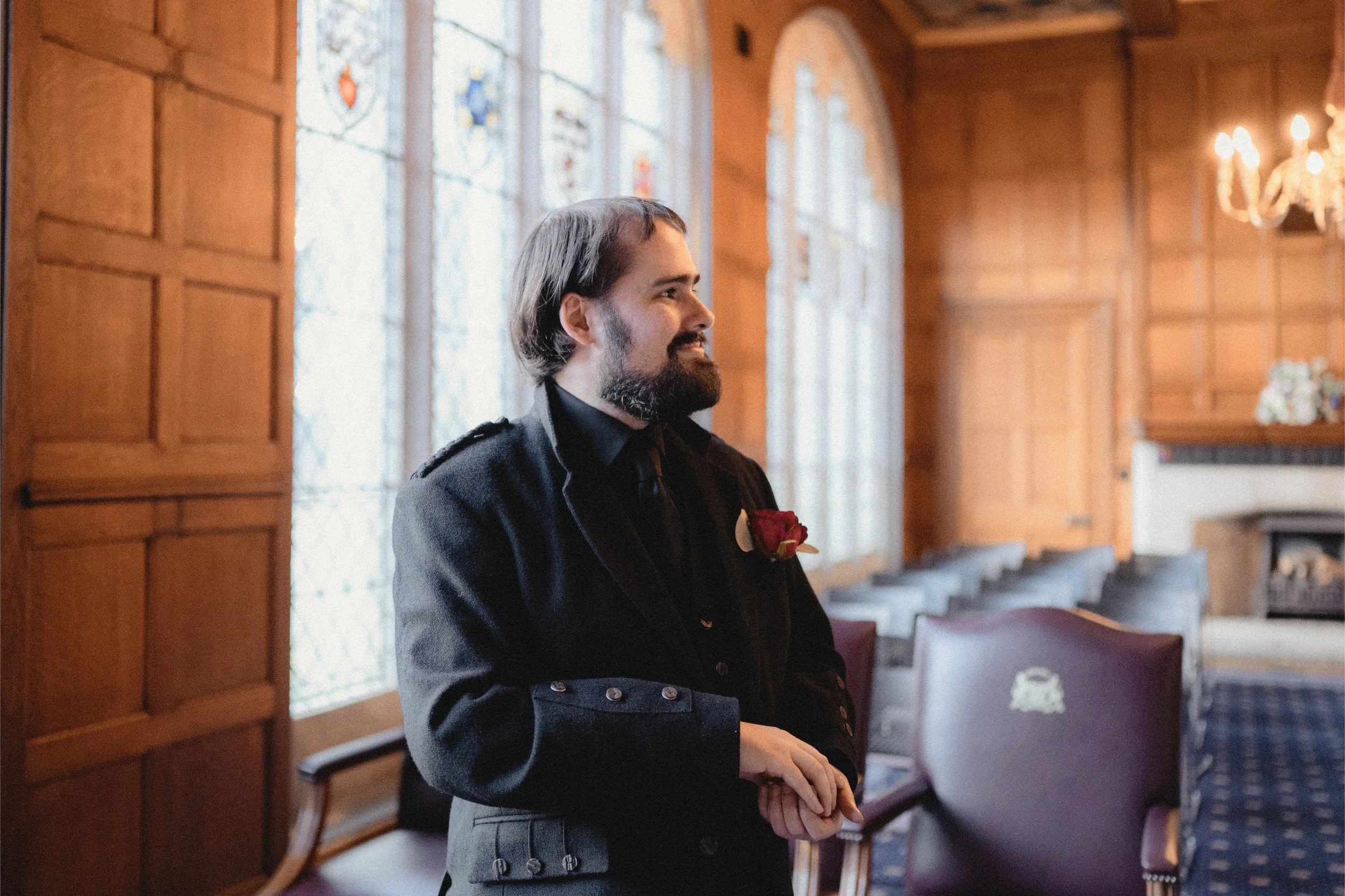 A man with a beard and long hair, dressed in a formal black military-style uniform with a red boutonniere, standing inside a wood-paneled room with stained glass windows and a chandelier.
