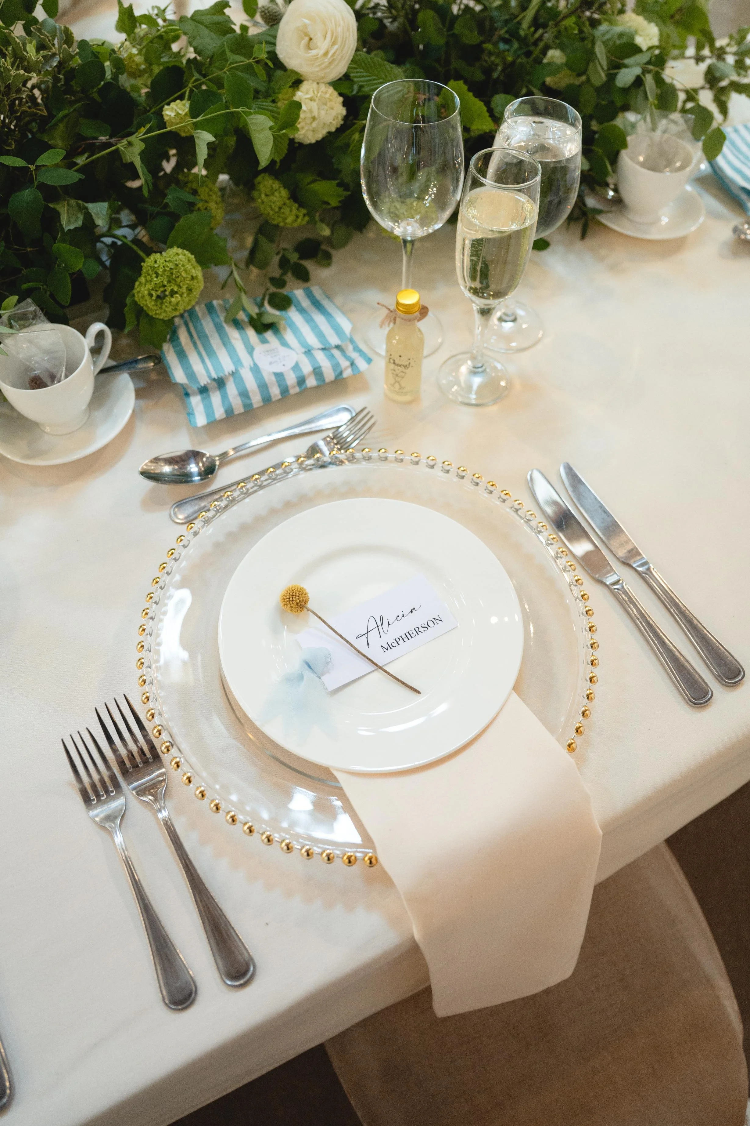 A formal table setting with a white tablecloth, a clear beaded charger plate, a smaller white plate with a name card that reads 'Alicia McPherson' and a dried yellow flower on top, and a beige napkin. Silverware includes two forks on the left and a k