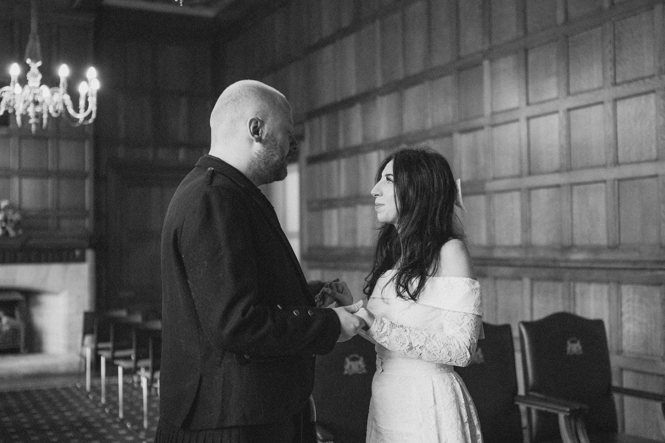 A couple holding hands in a wedding ceremony, with the groom dressed in a black jacket and the bride in a white lace off-the-shoulder wedding dress, inside a wood-paneled room with a chandelier.