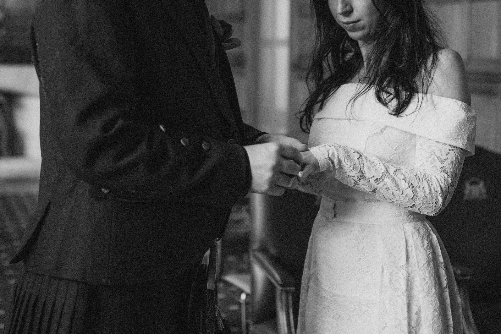 A black-and-white photo of a man and woman holding hands during a wedding ceremony. The woman is wearing a strapless lace wedding dress, and the man is dressed in a dark suit.