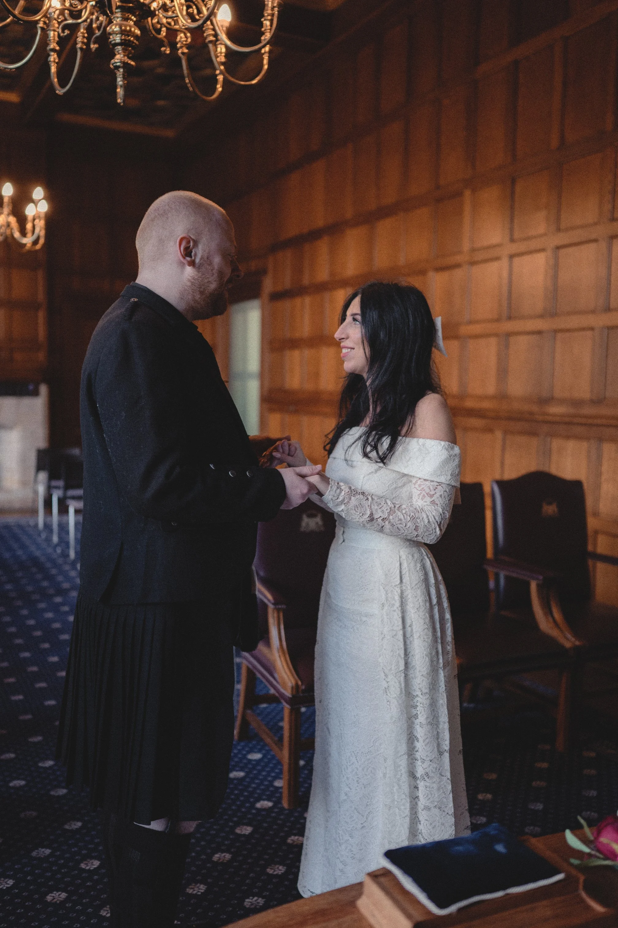 A couple is exchanging vows during a wedding ceremony in an elegant, wood-paneled room with chandeliers.