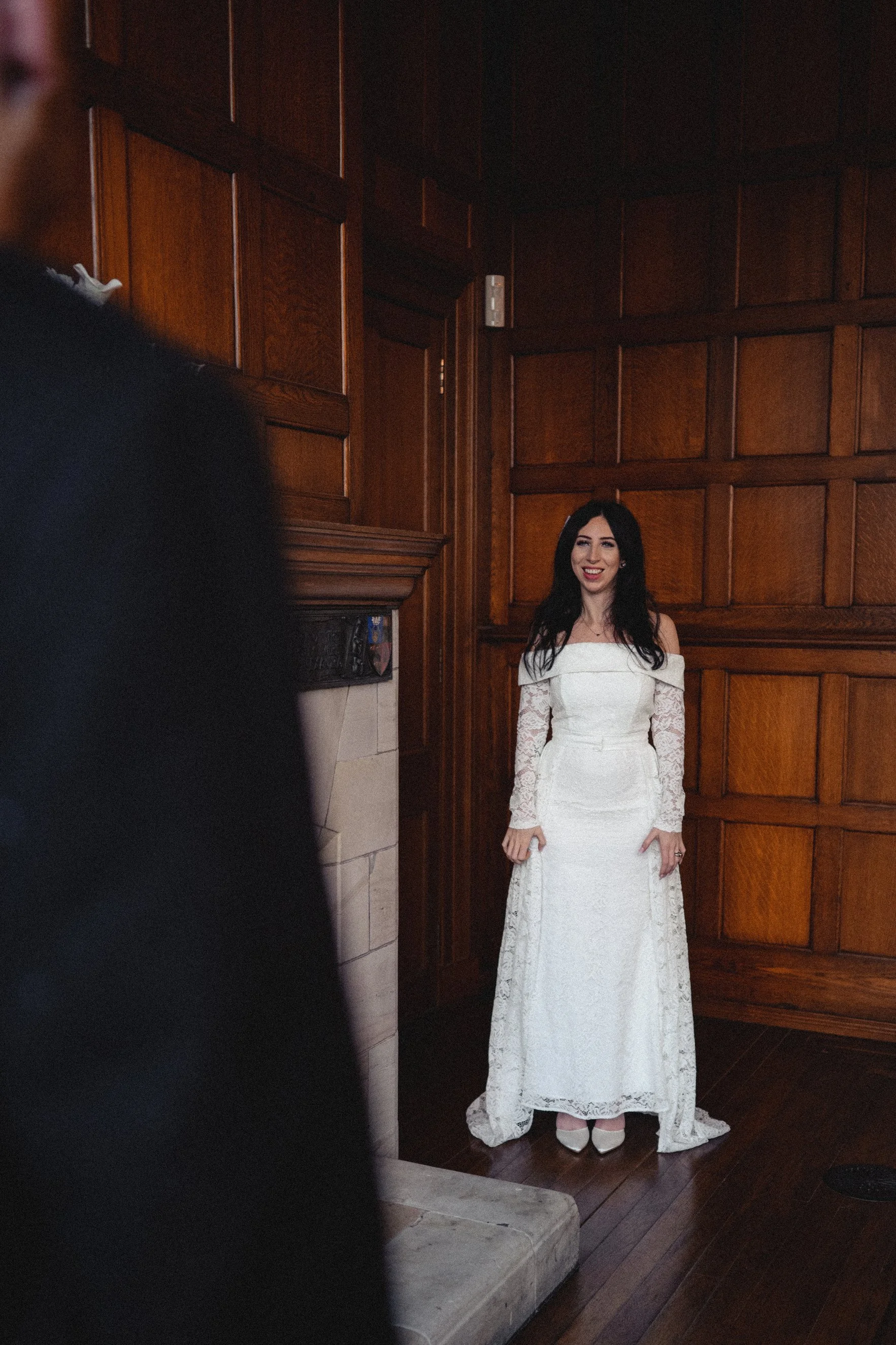 A woman in a white lace wedding dress standing indoors with wooden paneling, smiling at the camera.