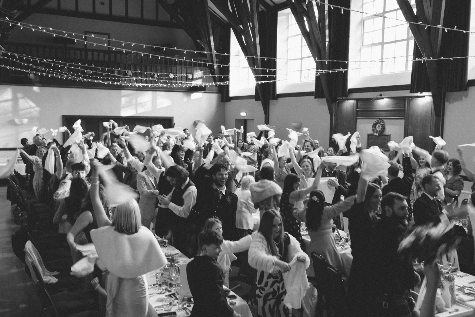 A large group of people gathered in a decorated hall, celebrating with some waving napkins and smiling, under string lights and high windows.