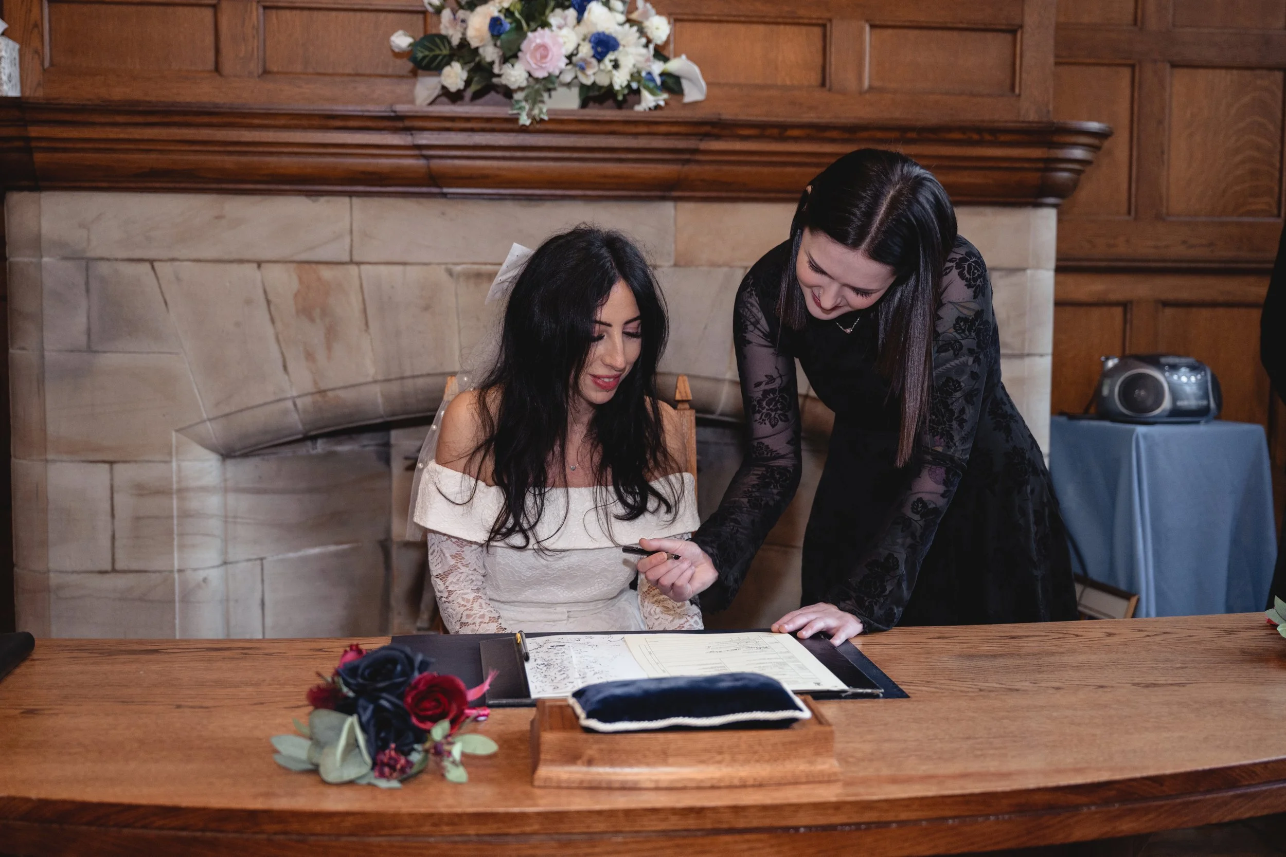 A woman in a white dress sits at a table with a bouquet of flowers nearby, while another woman in a black dress leans over and points at a document on the table during a wedding ceremony in a wood-paneled room with a stone fireplace and flower arrang