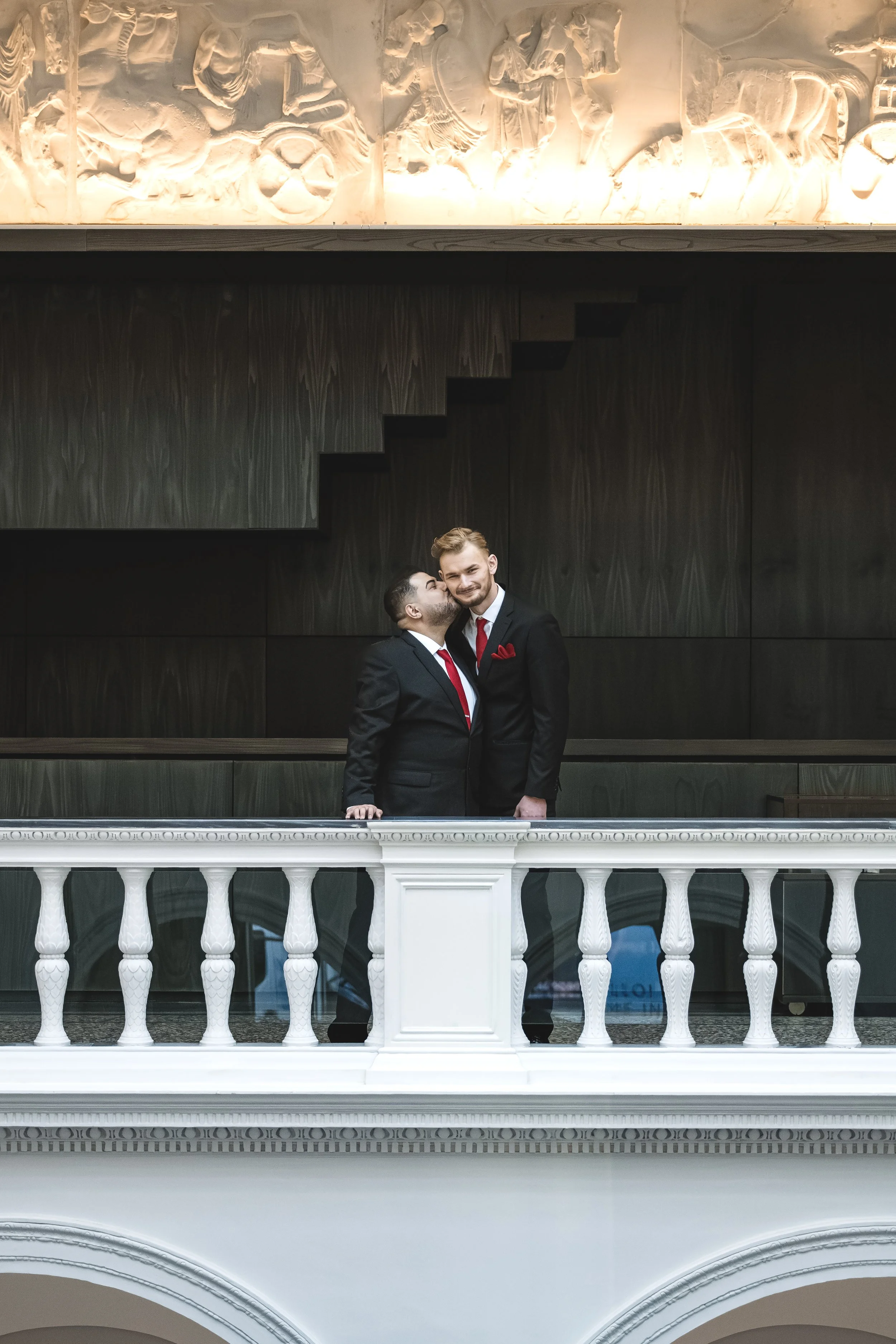 Two men in black suits and red ties, one kissing the other on the cheek in a formal setting with a white railing and dark wood paneling.