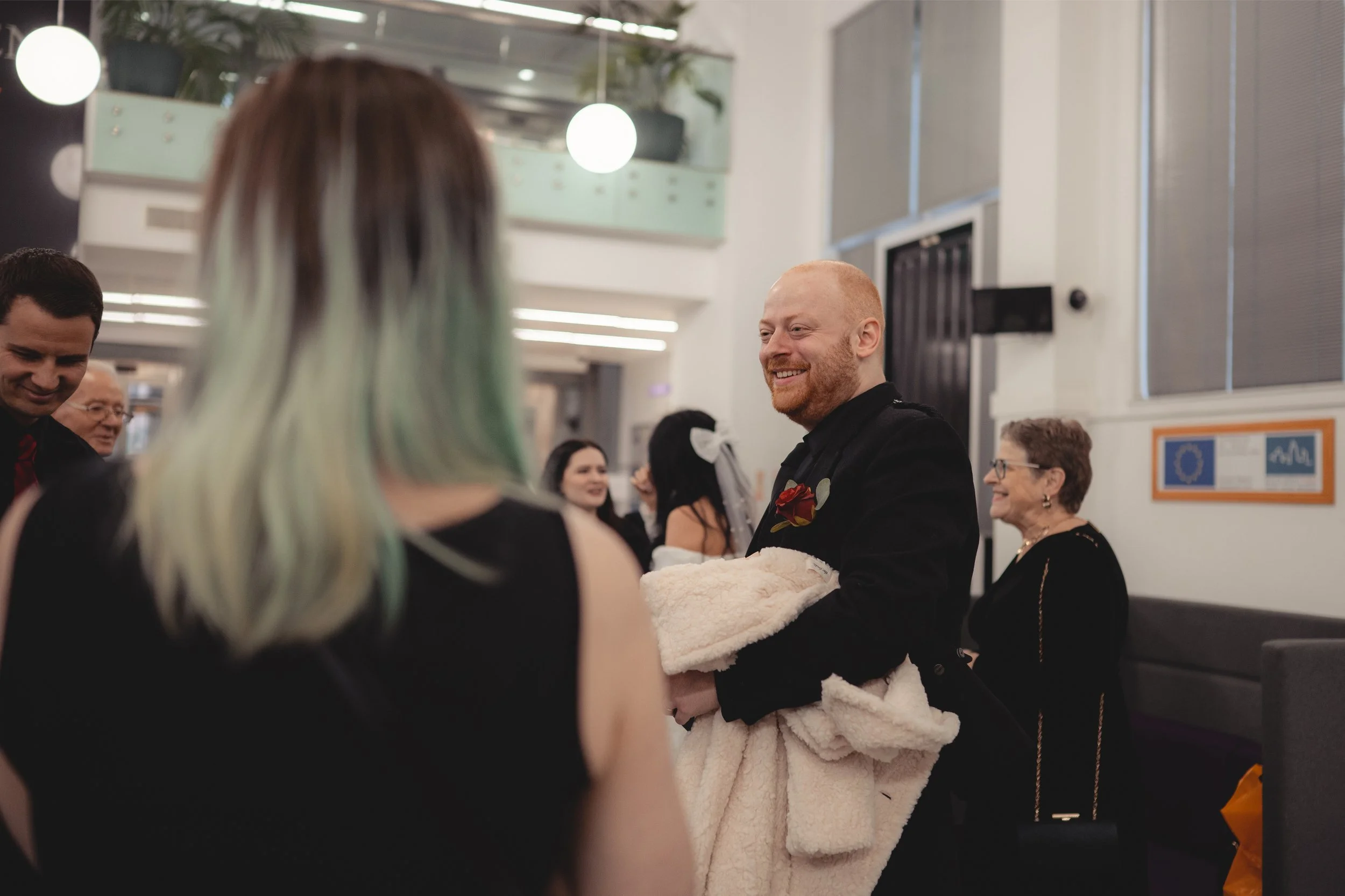 A man with a beard in black attire holding a blanket and smiling at a woman with multicolored hair at an indoor event with several other people around.