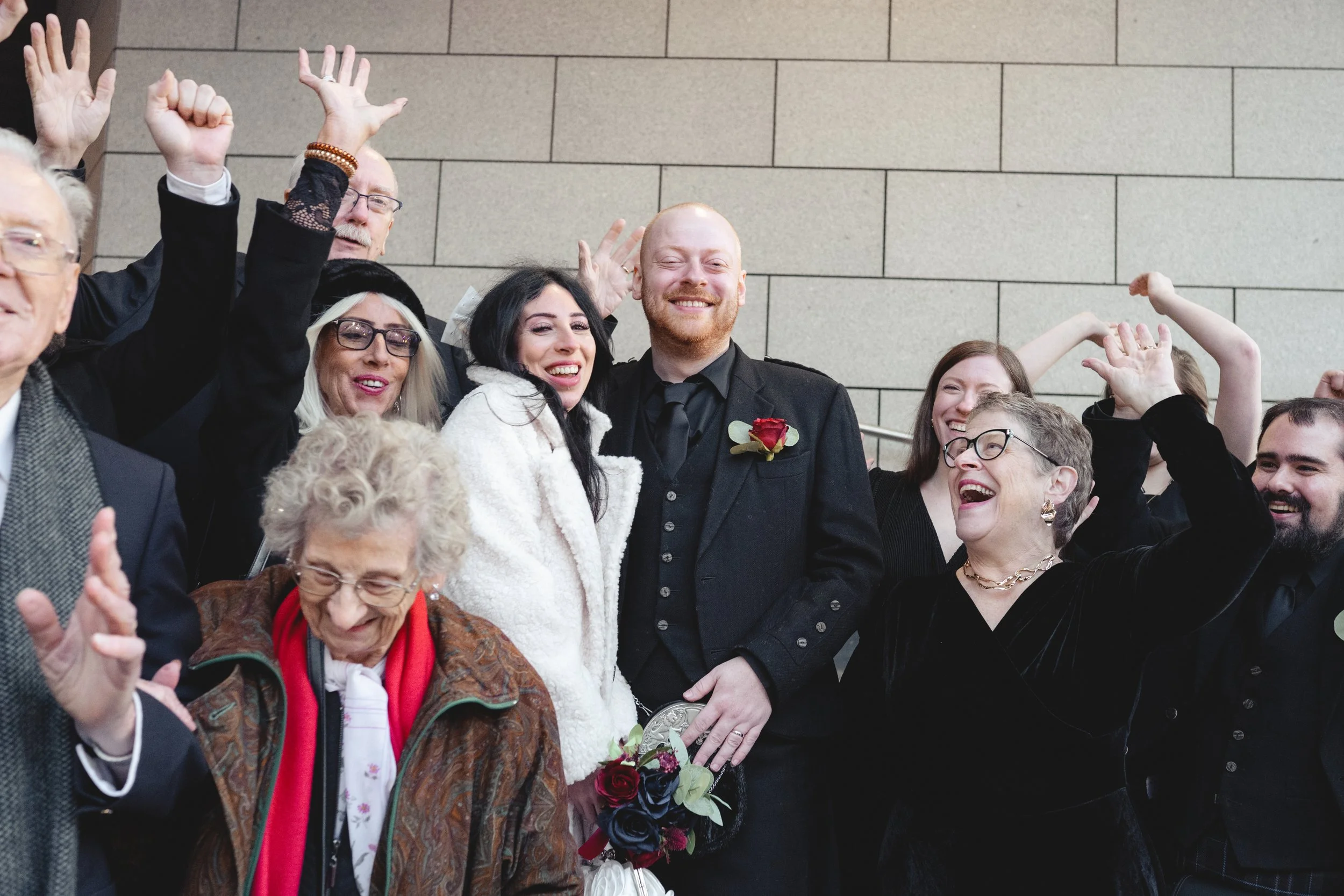 A group of people celebrating a wedding outdoors, with a bride and groom in the center, surrounded by friends and family smiling and raising their hands.