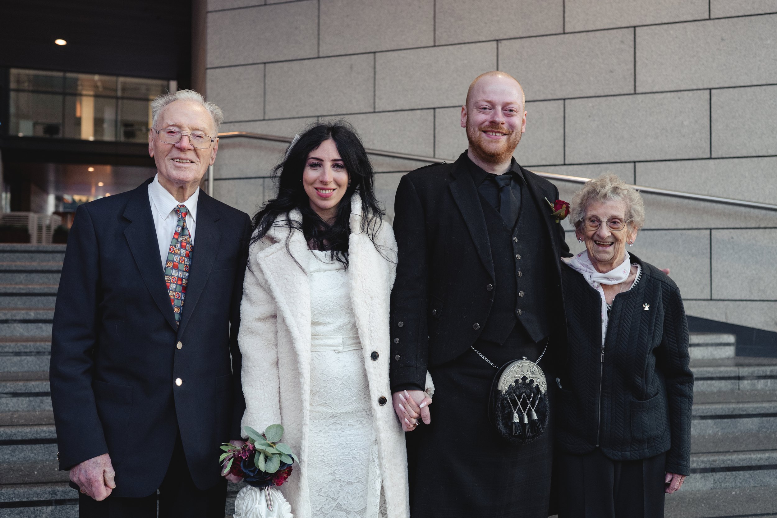 A group photo of four people standing outside a modern building. The group includes an elderly man in a suit, a woman in a white lace dress with a white coat, a man in black wedding attire with a red rose boutonniere, and an elderly woman in black at