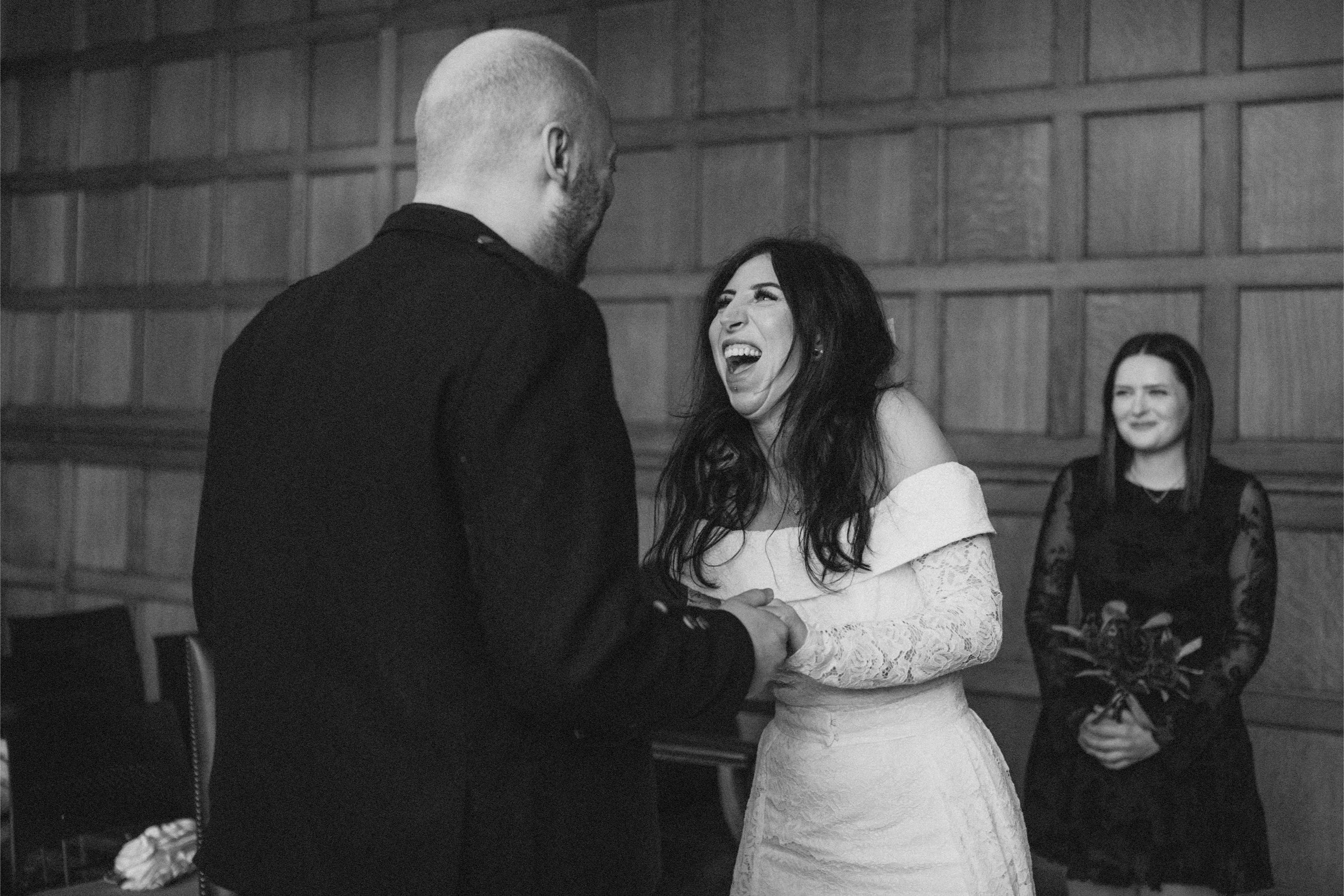 A woman in a white lace dress and a man in dark clothing hold hands and laugh at each other during a wedding ceremony in a wood-paneled room, with another woman in a black dress holding a bouquet smiling in the background.