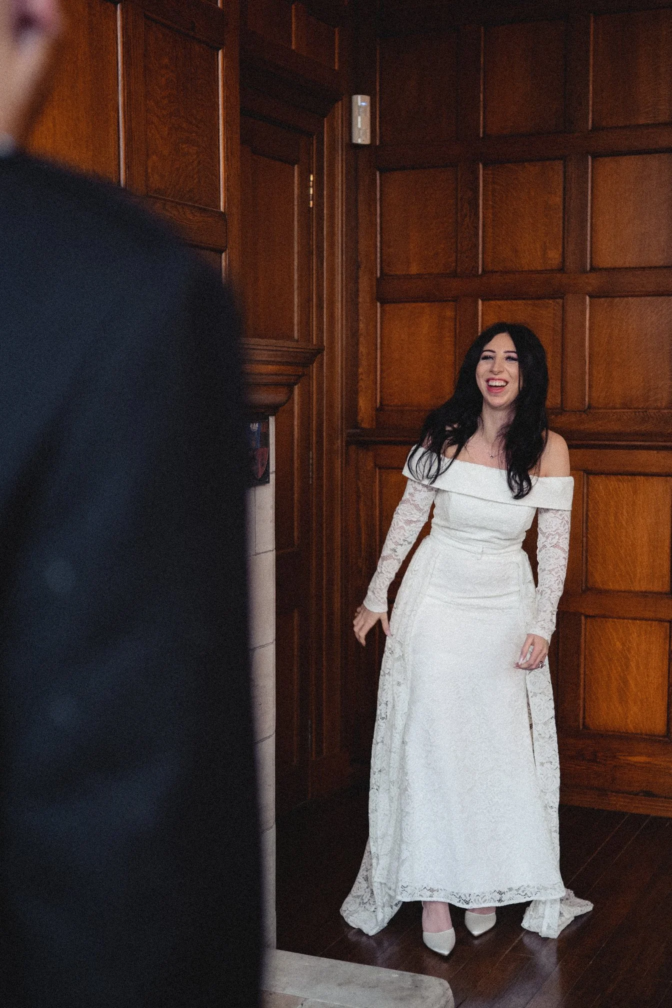 A woman in a white lace off-shoulder wedding dress with long sleeves is smiling and looking to her right, standing in a wooden-paneled room.