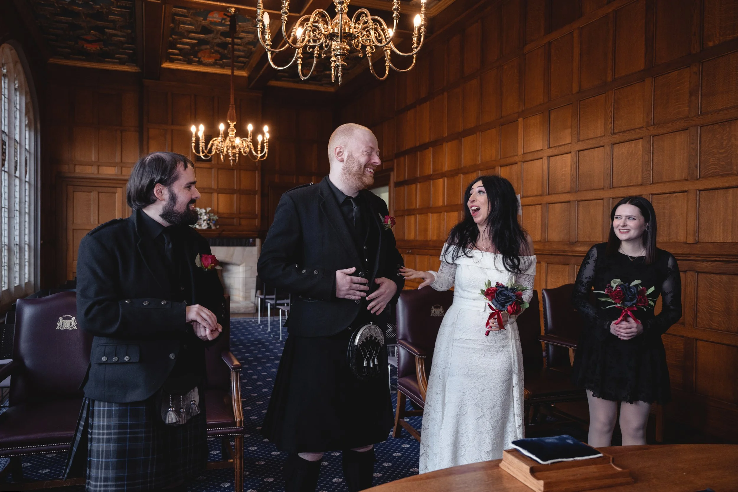 A wedding ceremony in a wood-paneled room with a chandelier, featuring a bride in a white dress holding a bouquet, a groom in a traditional kilt and black jacket, and two bridesmaids with bouquets, all smiling and laughing.