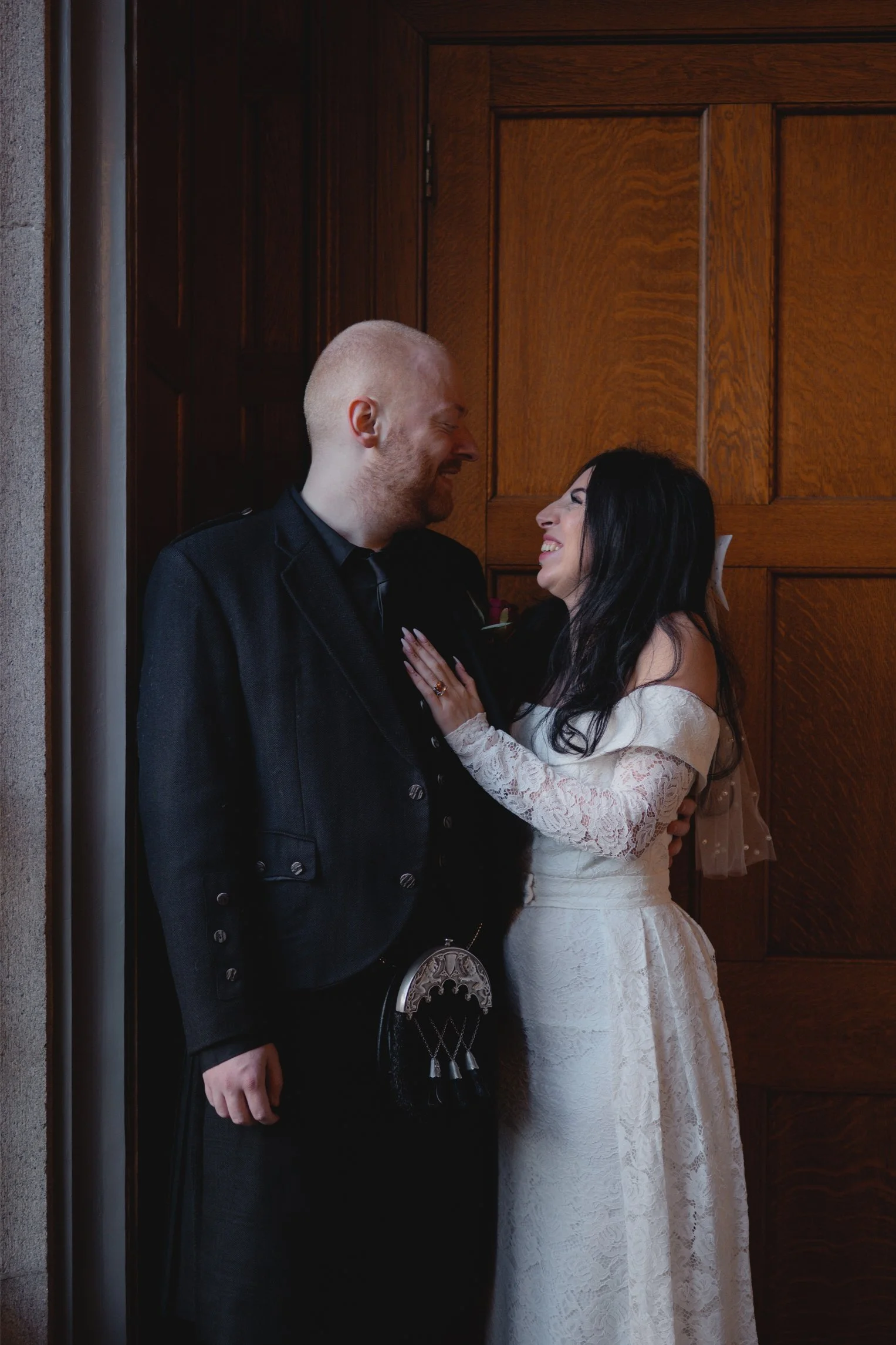 A couple in wedding attire sharing a joyful moment inside a wooden doorframe, with the groom in a traditional black kilt and jacket, and the bride in a white lace wedding dress.